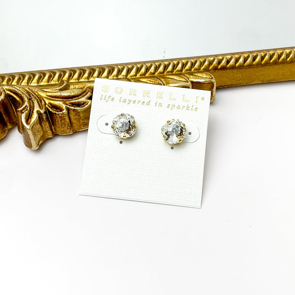 Pictured on a white cardstock is a pair of rounded square clear crystals with gold prongs. These earrings are pictured in front of a gold mirror on a white background. 