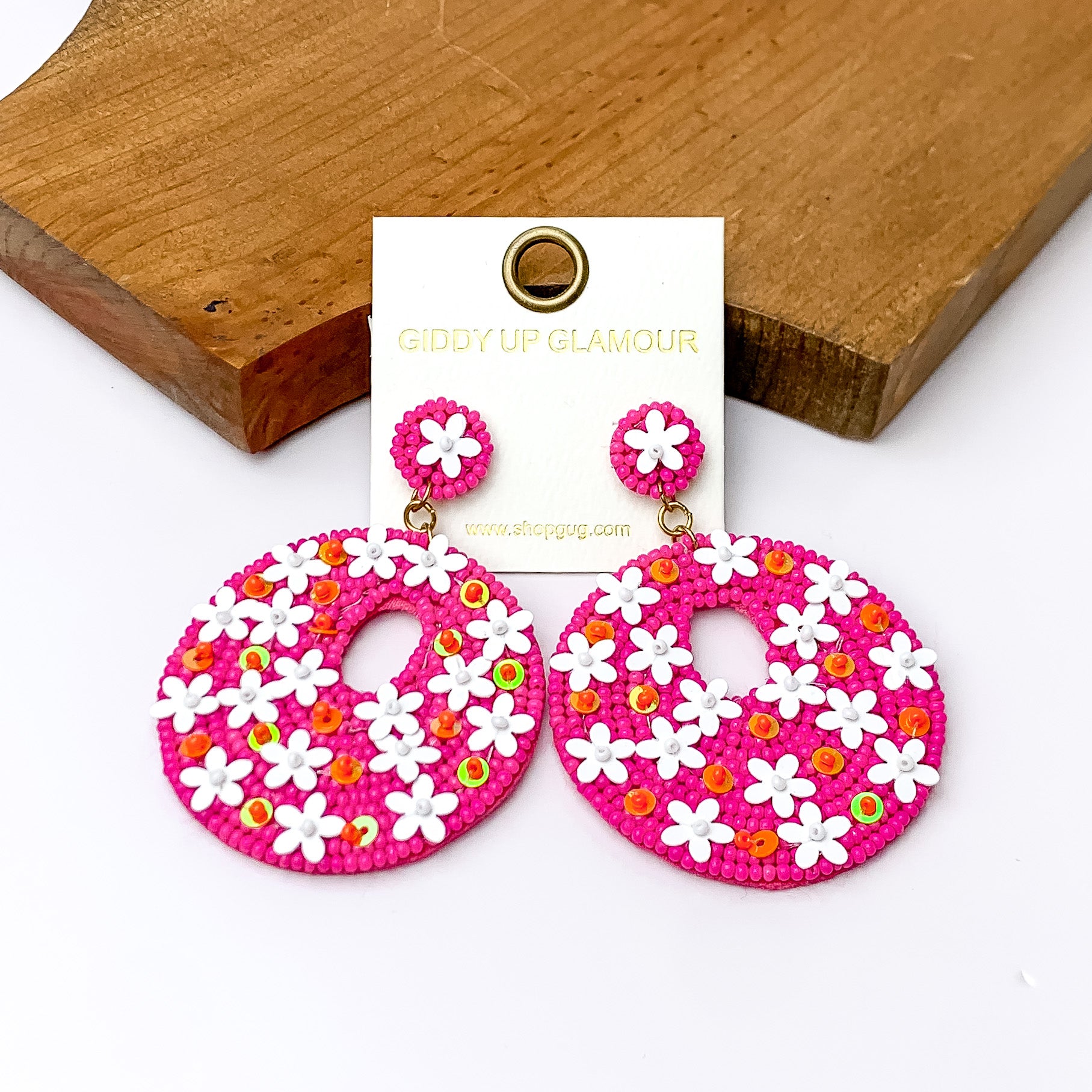 Hot pink beaded circular earrings with white and orange designs. Pictured on a white background with wood at the top.