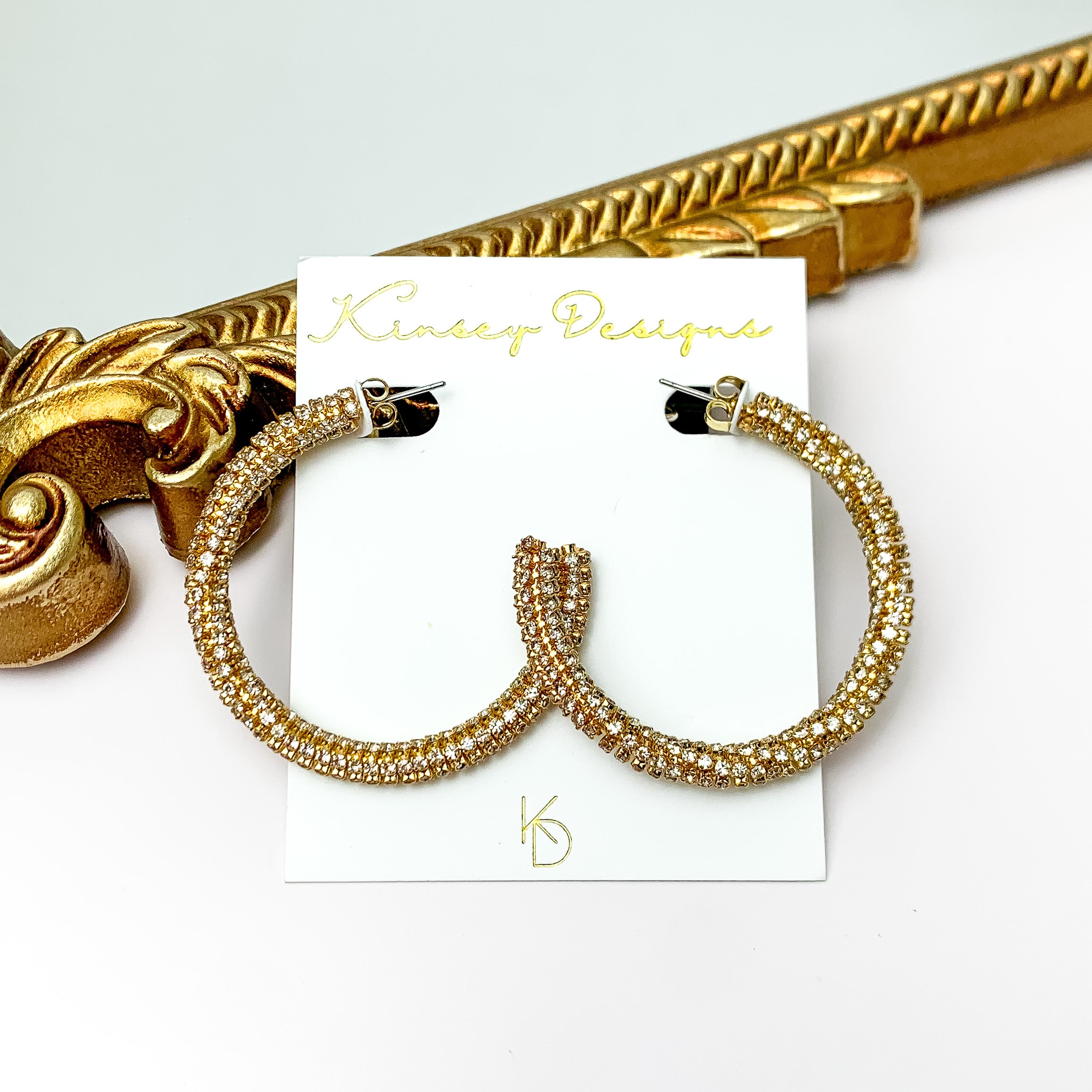 A pair of gold hoop earrings encrusted with clear crystals. These earrings are pictured in front of a gold mirror on a white background.  