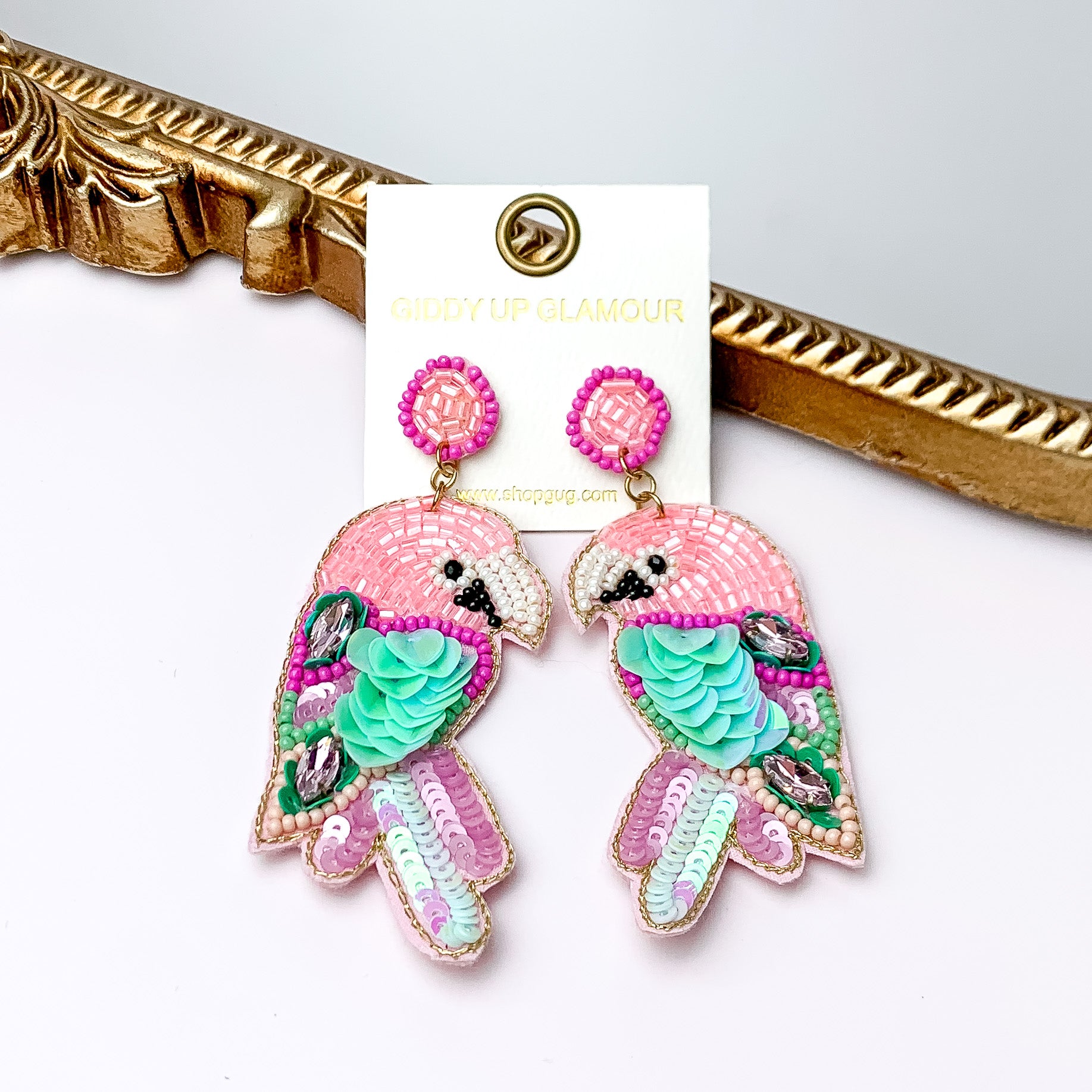 These parrot earrings are pink a little bit of turquoise, green, and purple beads. These earrings are pictured in front of a gold mirror on a white background.