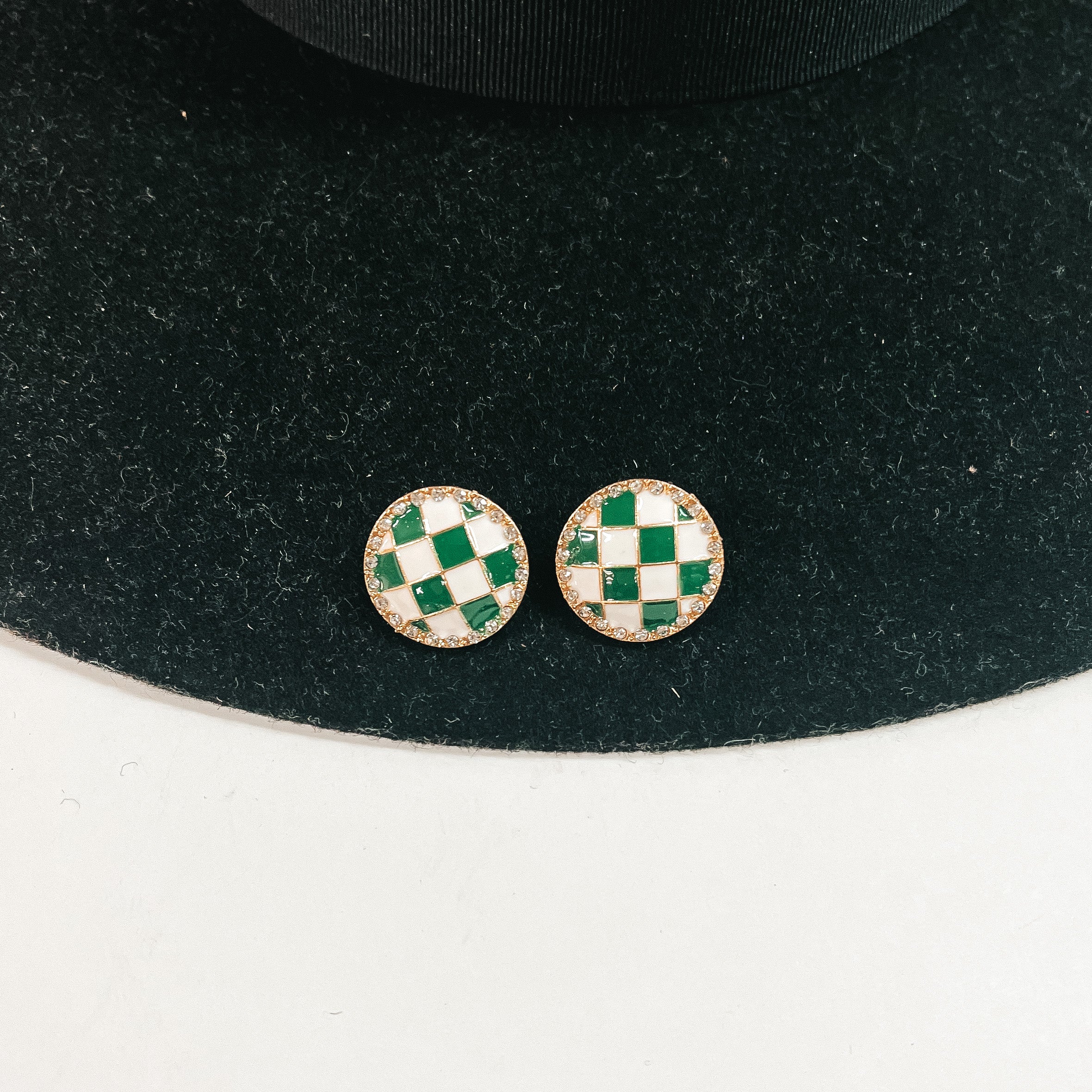 This is a pair of checkered patterned stud earrings in white/green in a gold  setting with clear crystals all around. This pair of earrings is laying on a  black felt hat brim and on a white background.