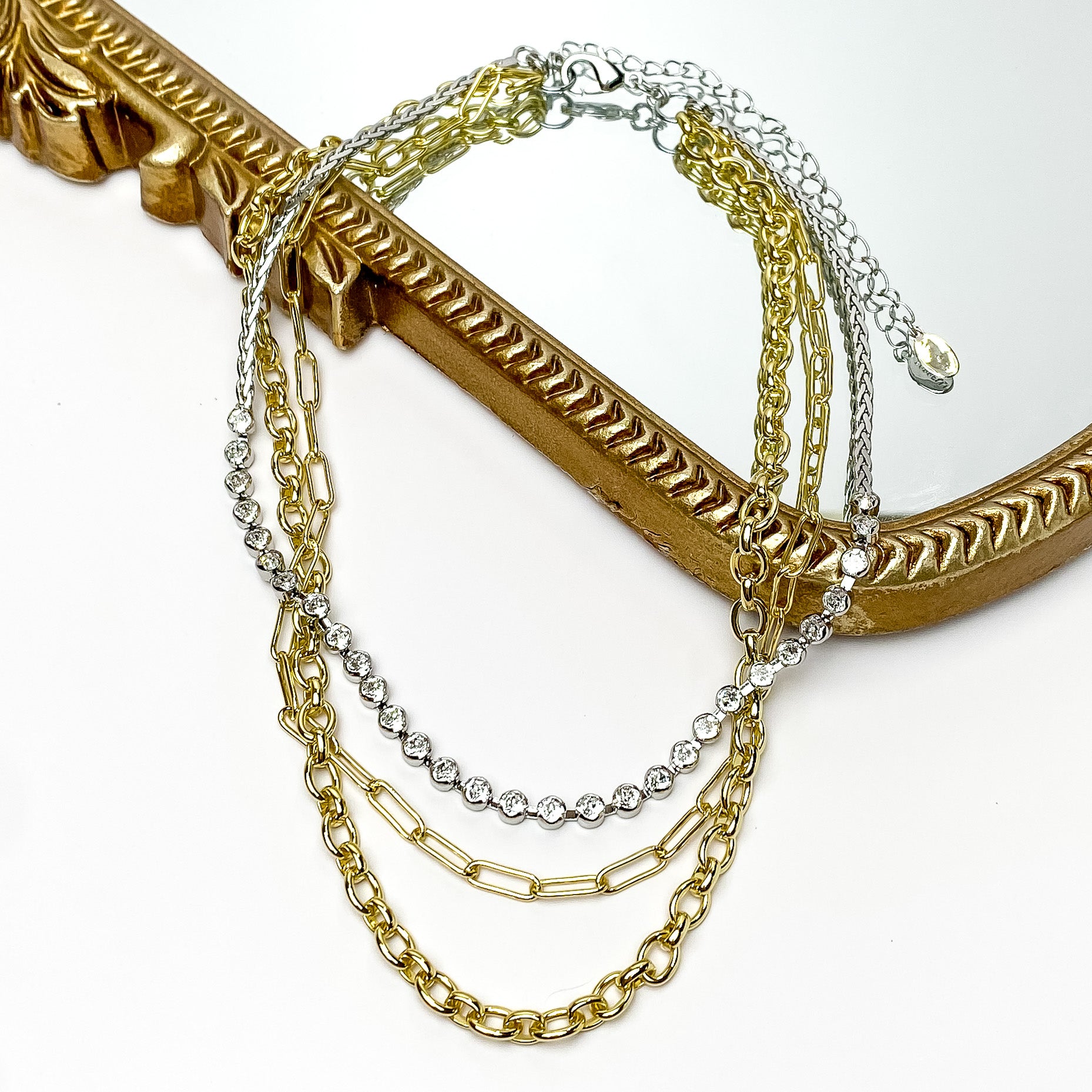 Pictured is a three strand necklace in mixed metal. There are two different chain strands in gold and the last strand is silver with round, clear crystals. This necklace is pictured partially laying on a gold mirror on a white background.    