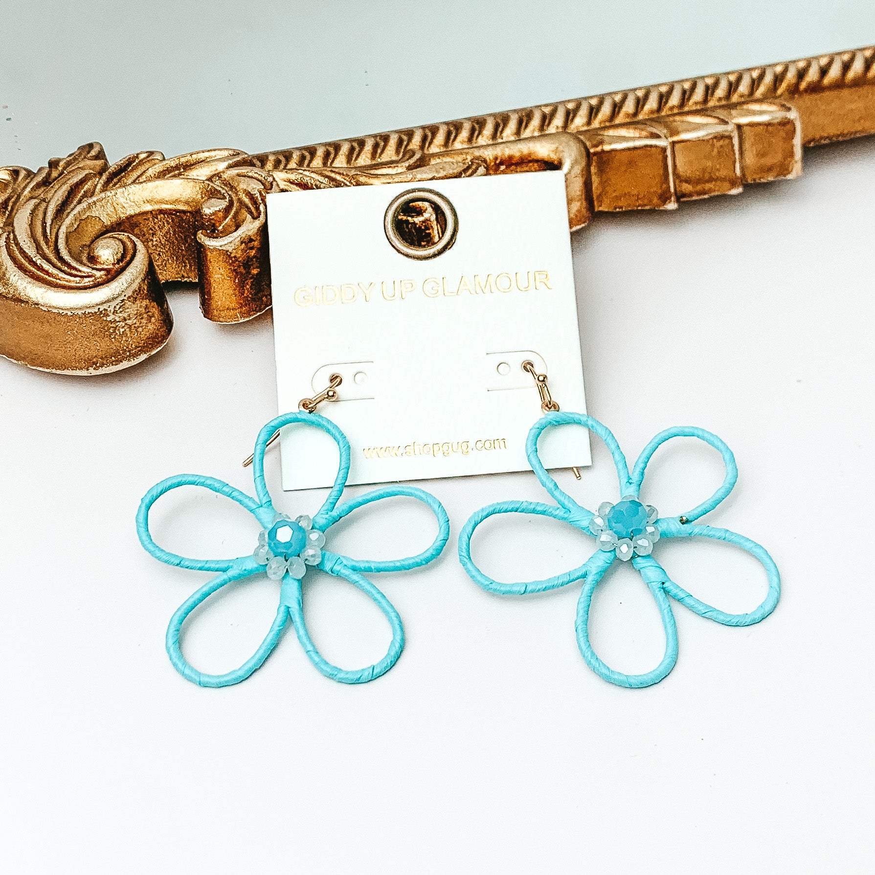 Pictured are gold fish hook earrings with a flower outline pendant. This pendant is pale turuoise and has center pale turquoise crystals. These earrings are pictured in front of a gold mirror on a white background. 