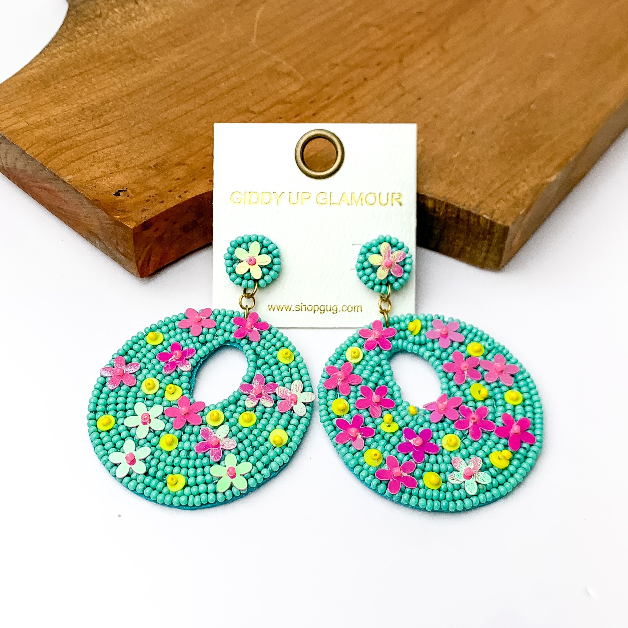 Turquoise beaded drop circular earrings with floral designs. Pictured on a white background with a wood piece at top. 