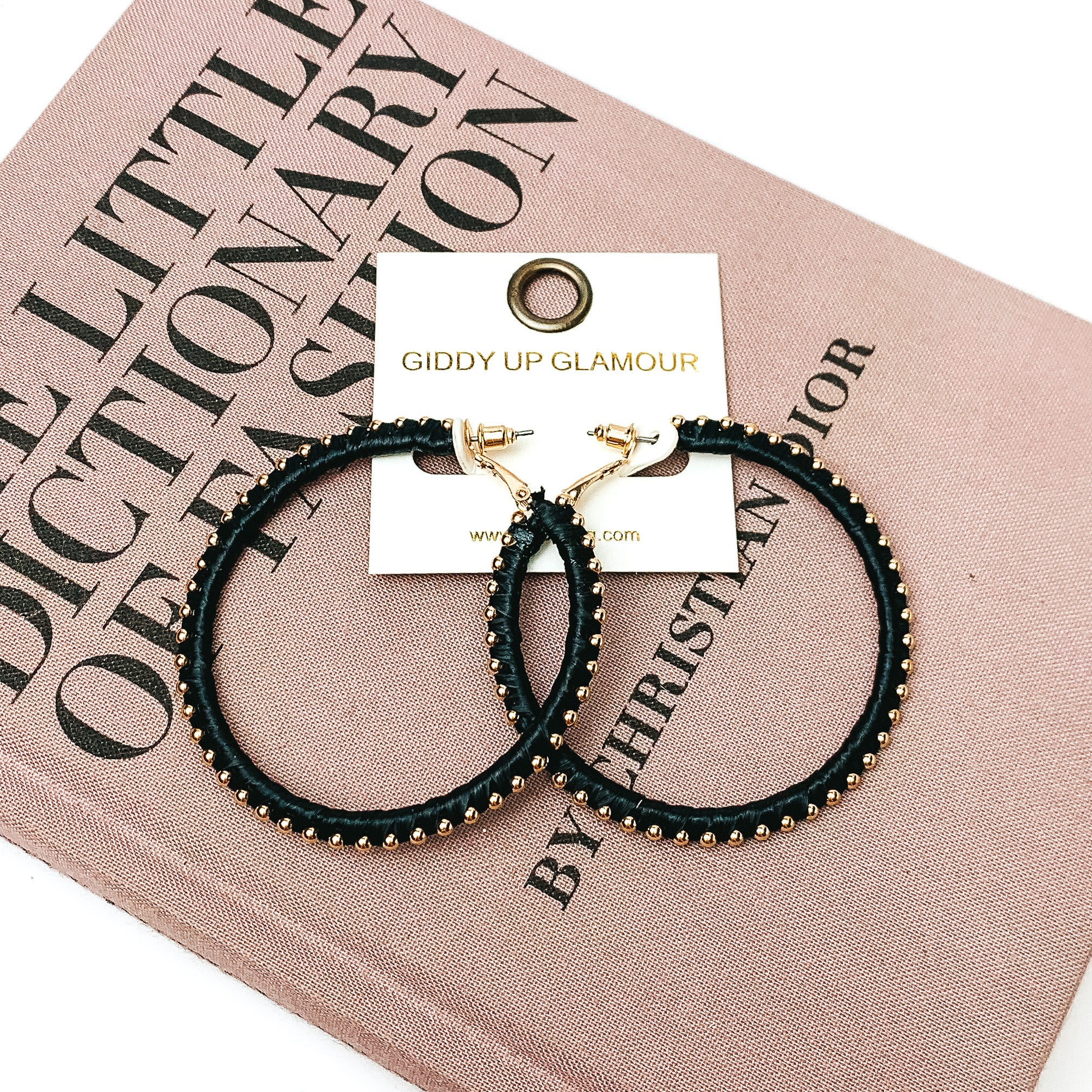 Pictured are circle black hoop earrings with gold beades around it. They are pictured with a pink fashion journal on a white background.