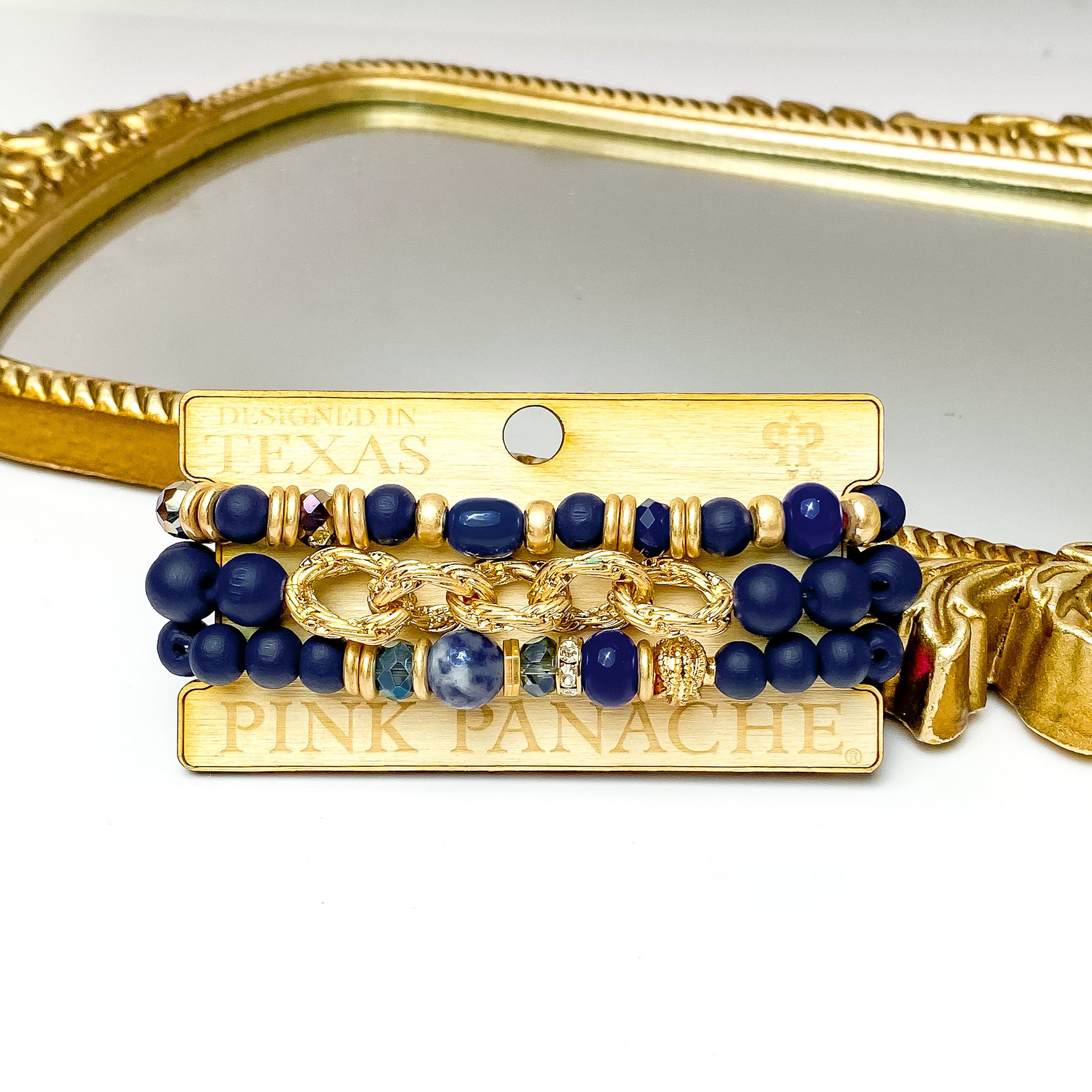 Set of three navy and gold bead bracelets in different sizes.These bracelets also include a gold chain segment and different types of navy and gold beads on one bracelet. These braceets are pictured on a Pink Panache wood holder in front of a gold mirror and on a white background. 