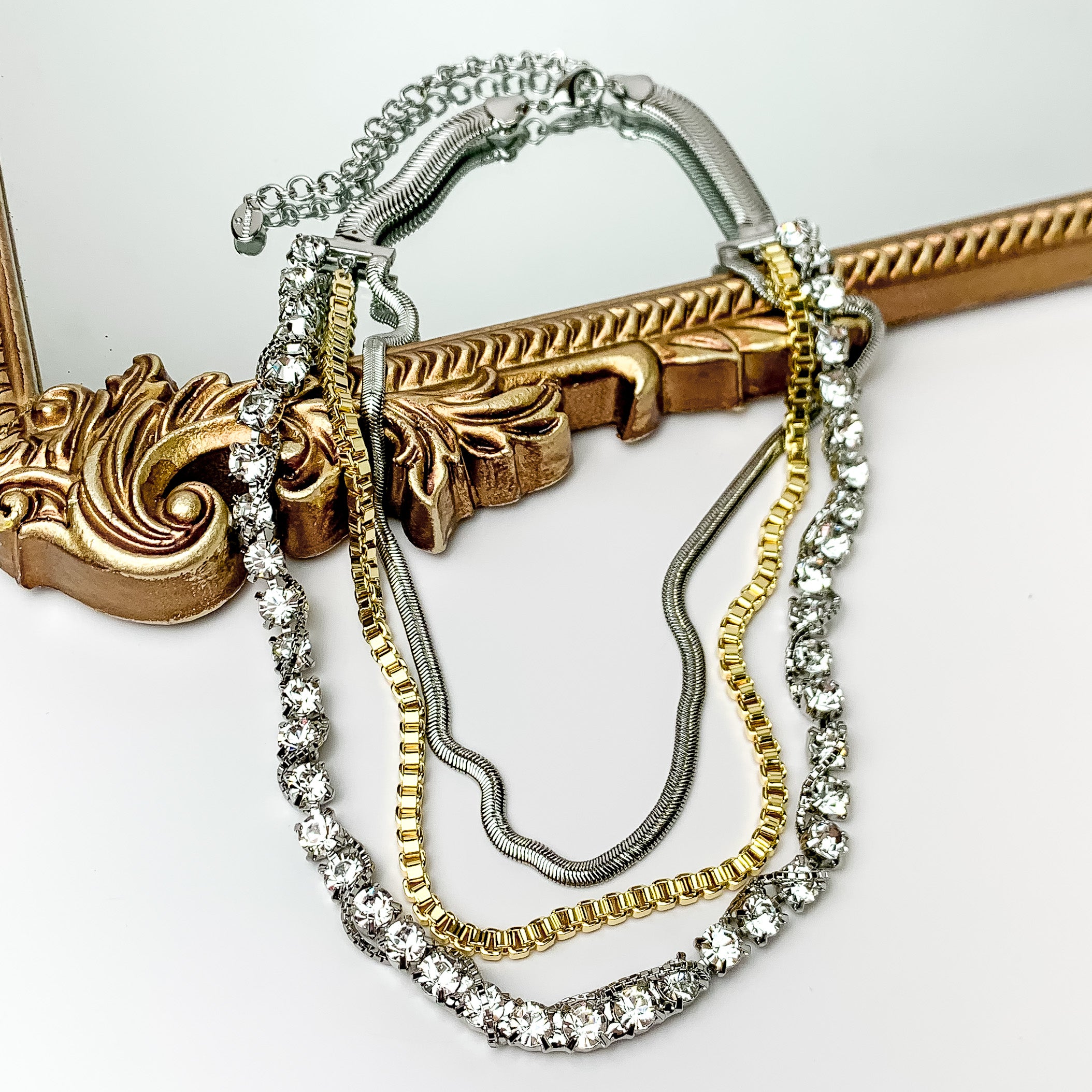 Pictured is a three strnad necklace. This necklace includes a clear crystal strand with a silver chain wrapped around the crystals, a gold box chain strand, and a silver herringbone chain strand. This necklace is pictured partially on a gold mirror on a white background. 