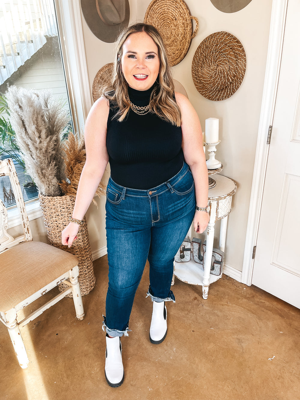Model is wearing a blac, ribbed, turtleneck, tank top bodysuit with dark wash skinny jeans. The jeans are rolled up with a raw hem, she is also wearing white booties and gold jewelry. Such as gold bracelets and necklaces.