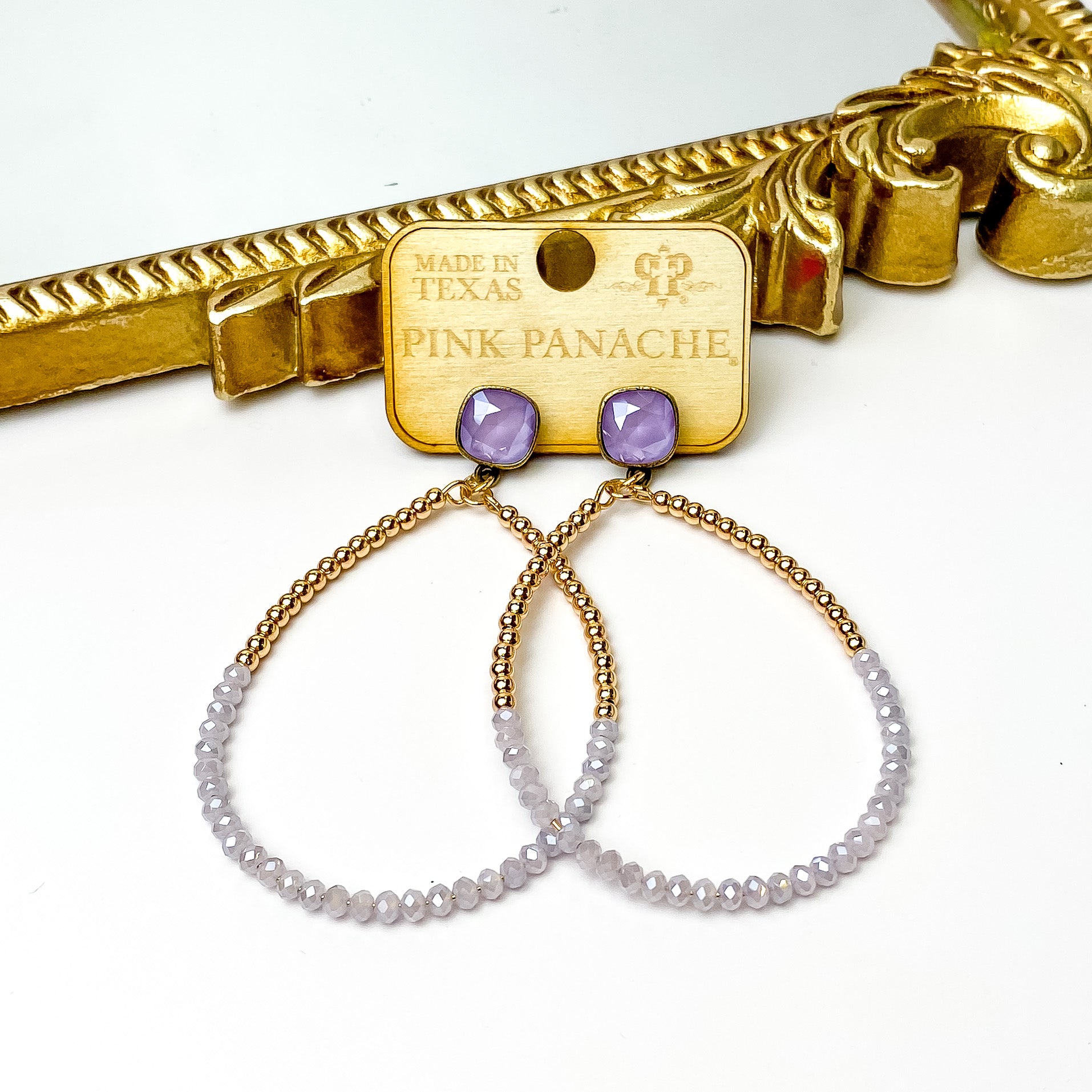 Lilac cushion cut crystal stud earrings with a teardrop pendant. The pendant is half gold beads and half lavender crystal beads. These earrings are pictured on a wood earrings holder in front of a gold mirror on a white background. 