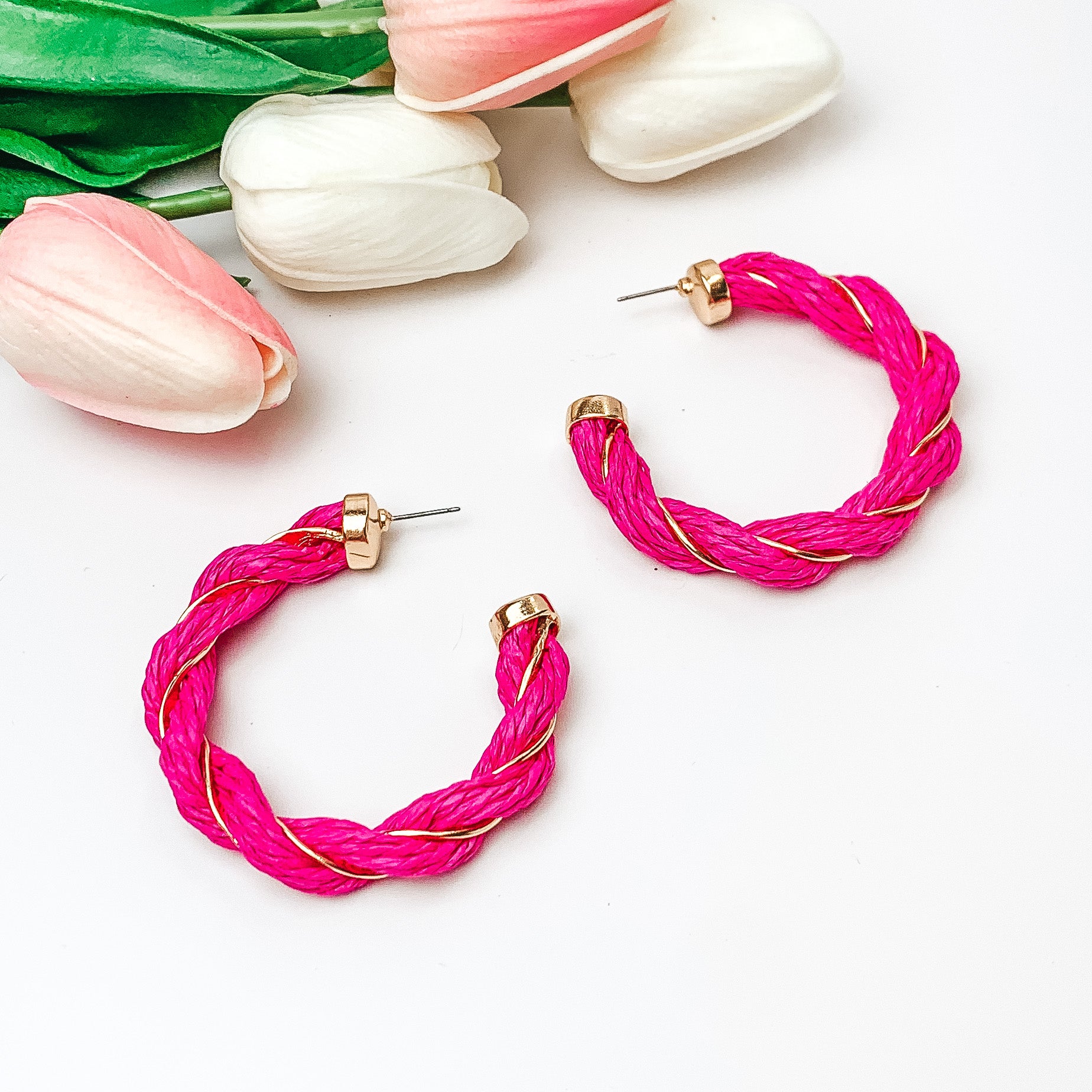 Pictured are fuchsia raffia twisted hoop earrings with gold detailing.  They are pictured with pink and white tulips on a white background.