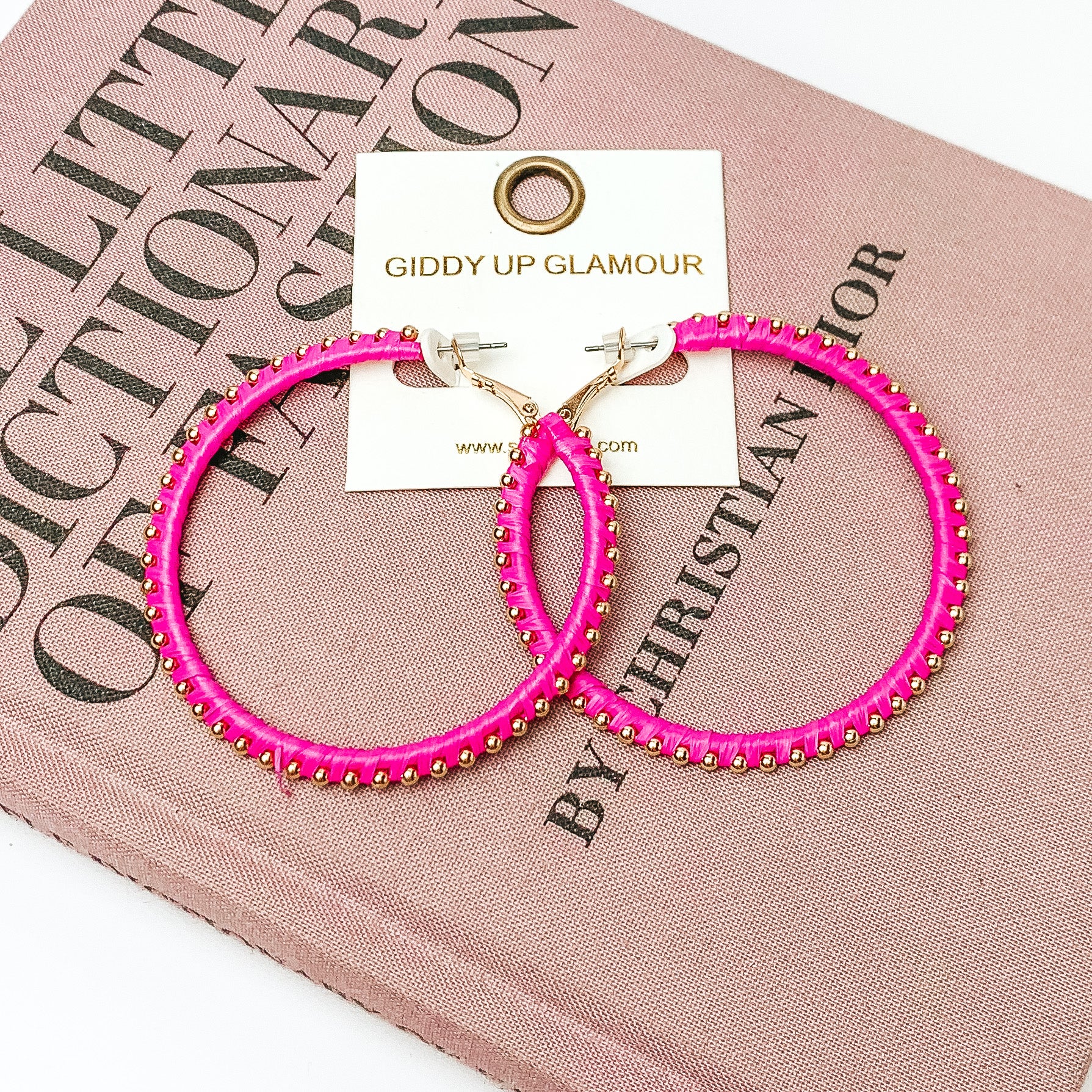 Pictured are circle hot pink hoop earrings with gold beads around it. They are pictured with a pink fashion journal on a white background.