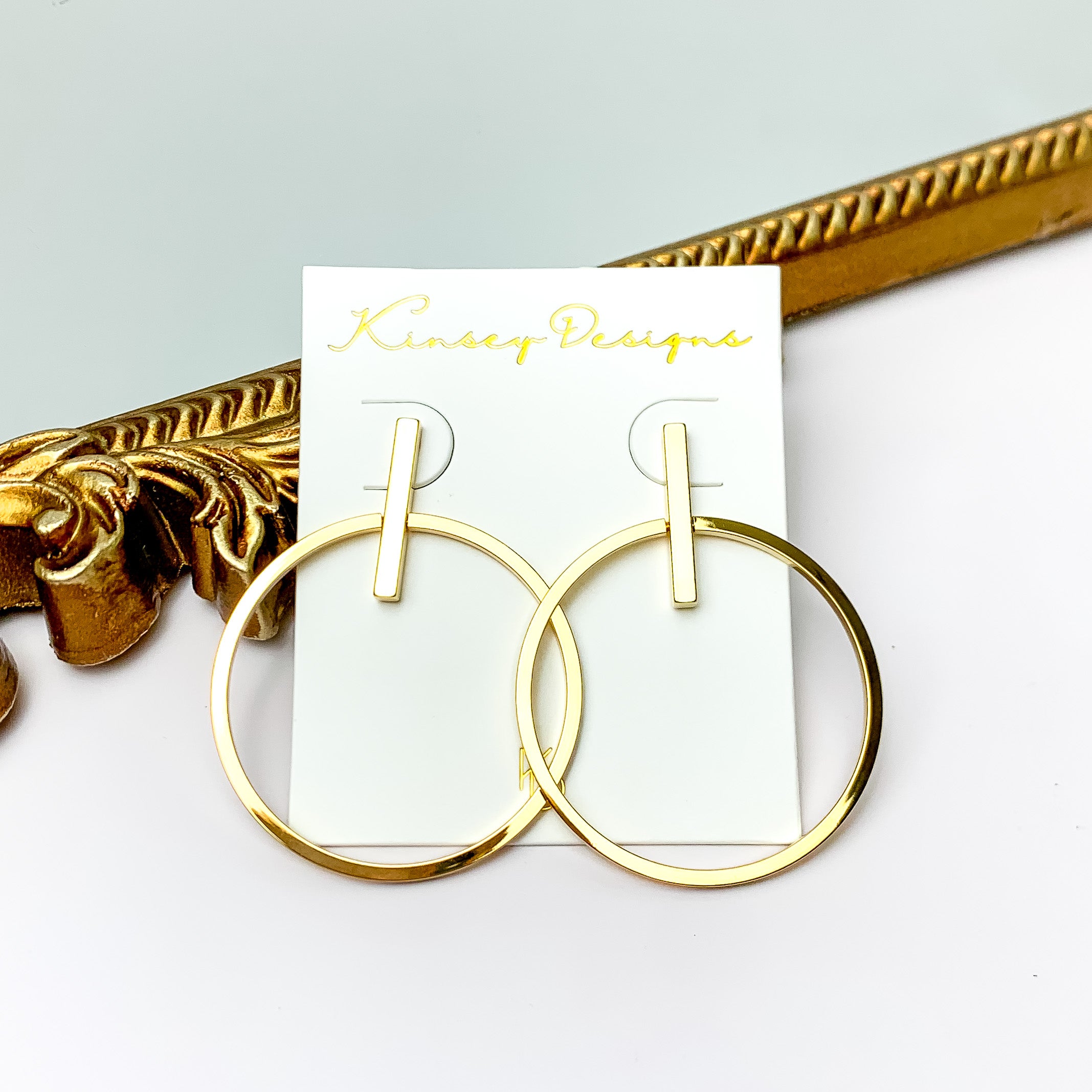 A pair of gold hoop earrings with a rectangle bar post back. These earrings are pictured in front of a gold mirror on a white background.  