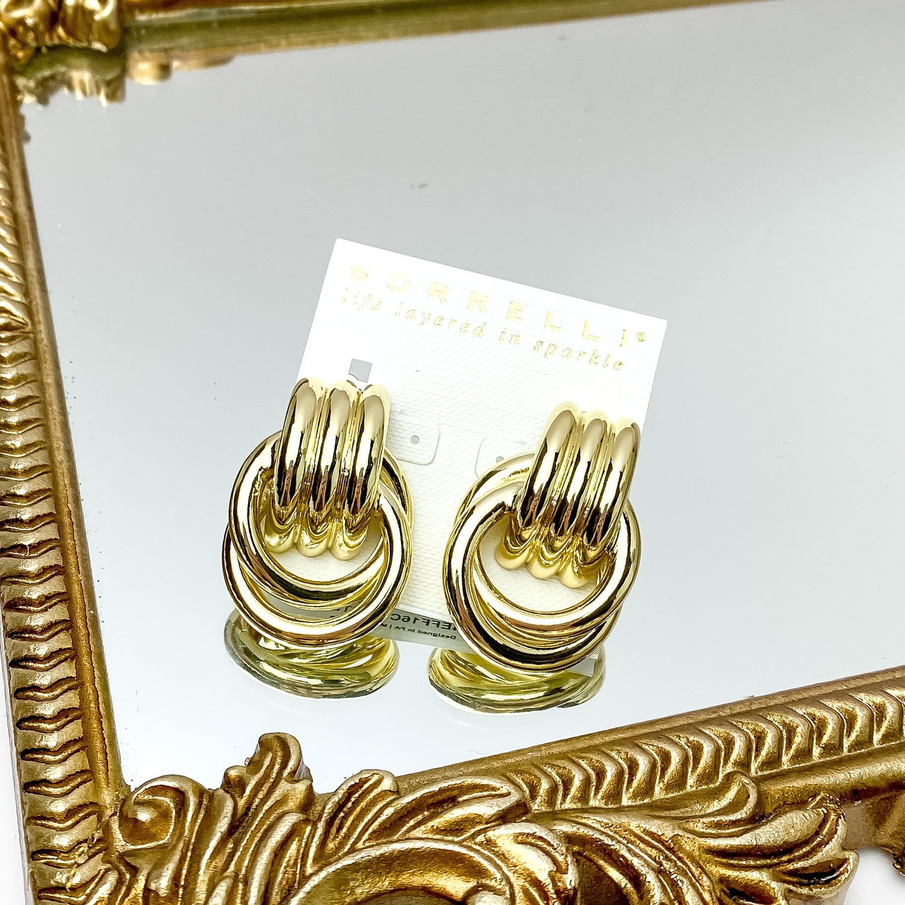 Two chunky gold earrings. These earrings feature links fused together and wrapped around eachother and pictured on a white cardstock. These earrings are pictured on a gold mirror on a white background. 