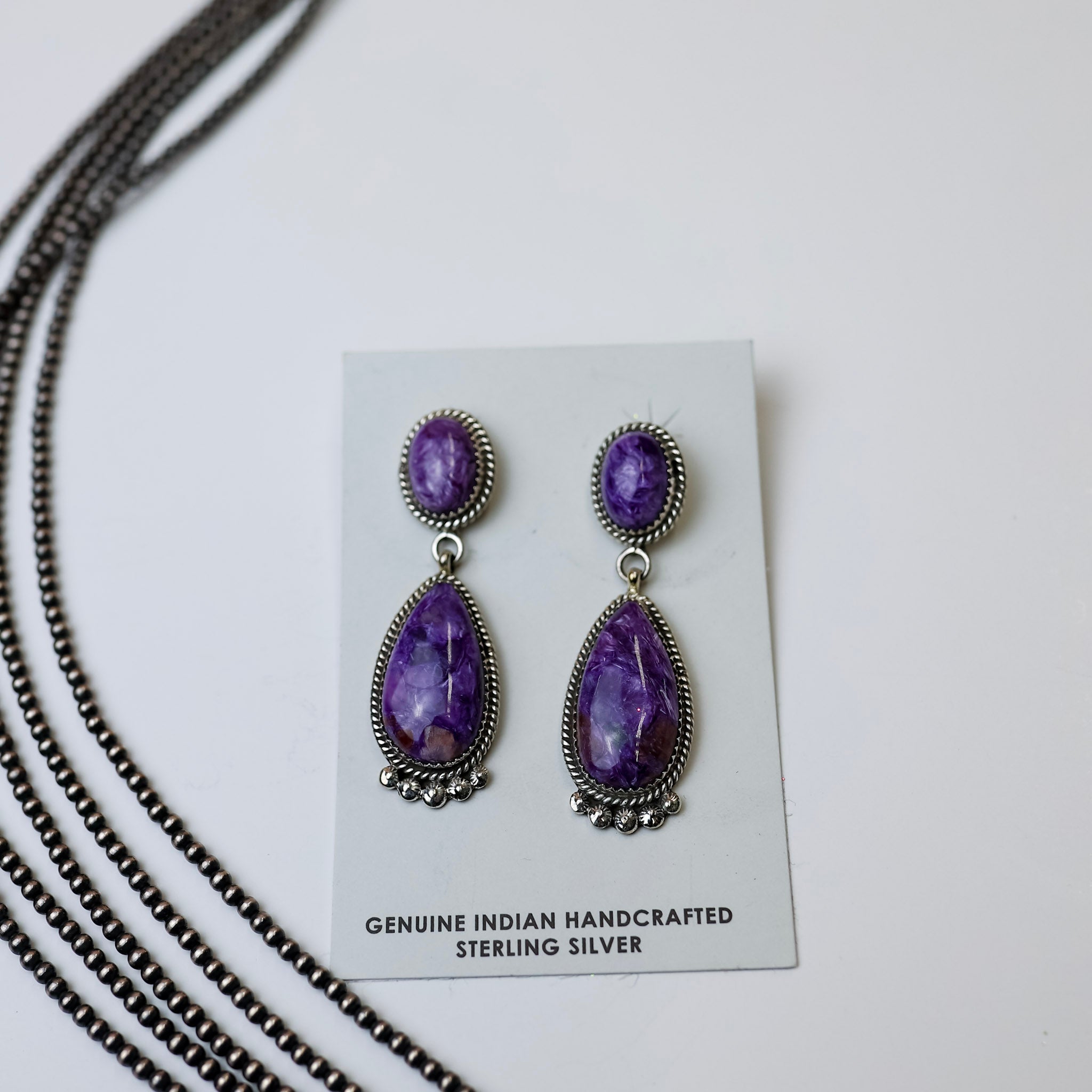Elouise Kee Navajo Handmade Sterling Silver & Charoite Drop Earrings are centered in the middle of the picture on a white background. Navajo pearls are on the left side of the picture. 