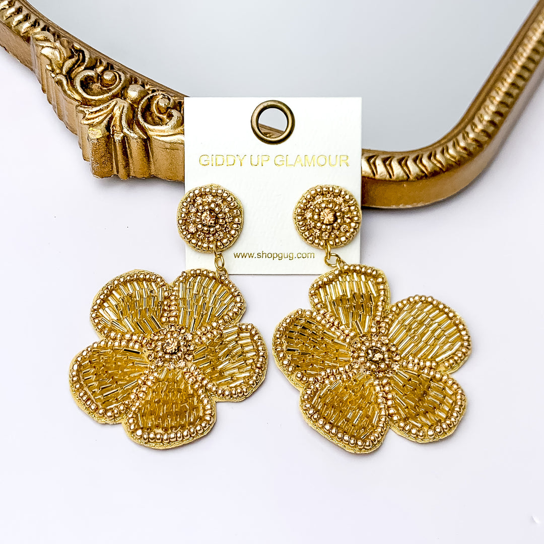 There is hanging beaded flower pendant from the gold stud earrings. The flower pendants are gold with  gold outline and detailing. These earrings are pictured in front of a gold mirror on a white background.