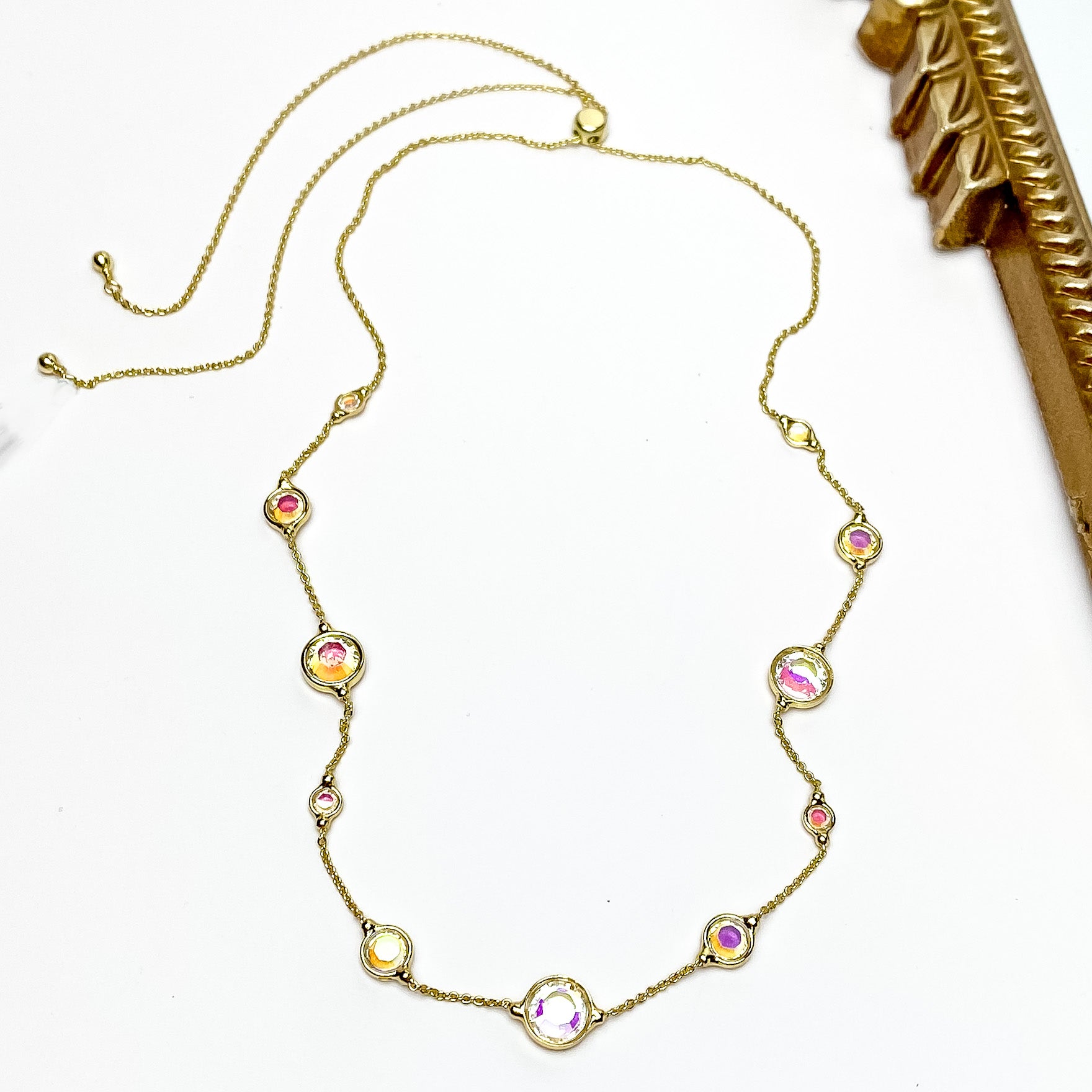 Pictured is a thin, gold chain with spaced out circle ab crystals. These crystals also come in different sizes. This necklace is pictured on a white background with a gold mirror to the right of the necklace.    