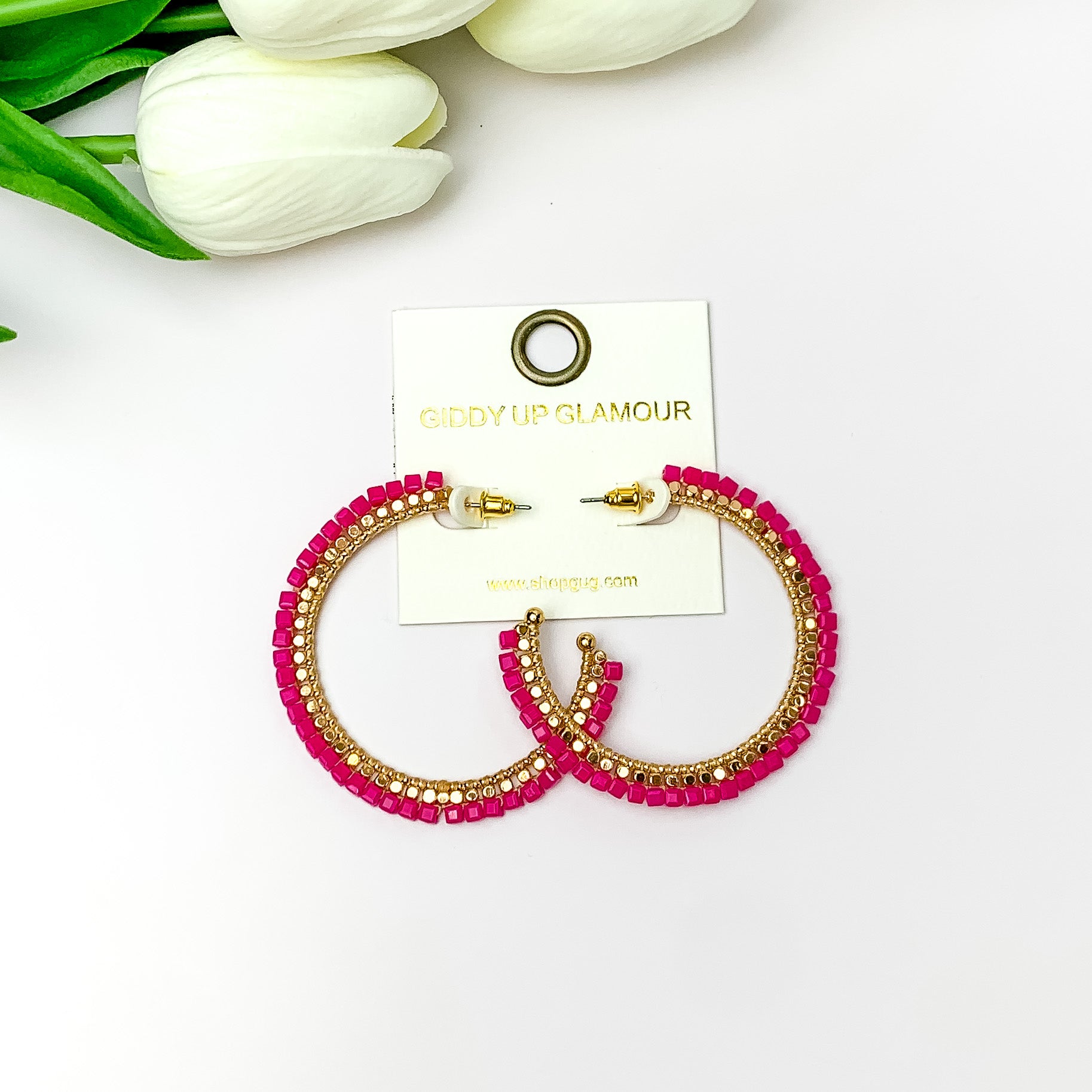 Pictured are circle gold toned hoop earrings with gold beads around it and hot pink crystal outline. They are pictured with white flowers on a white background.