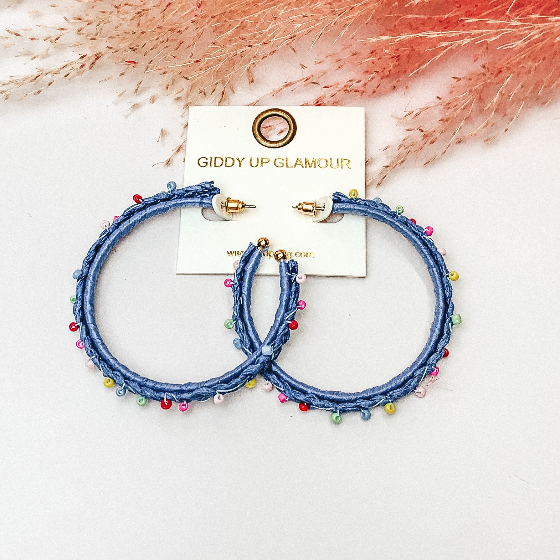 Pictured are raffia braided hoop earrings in blue with colorful beads. They are pictured with a pink feather on a white background.