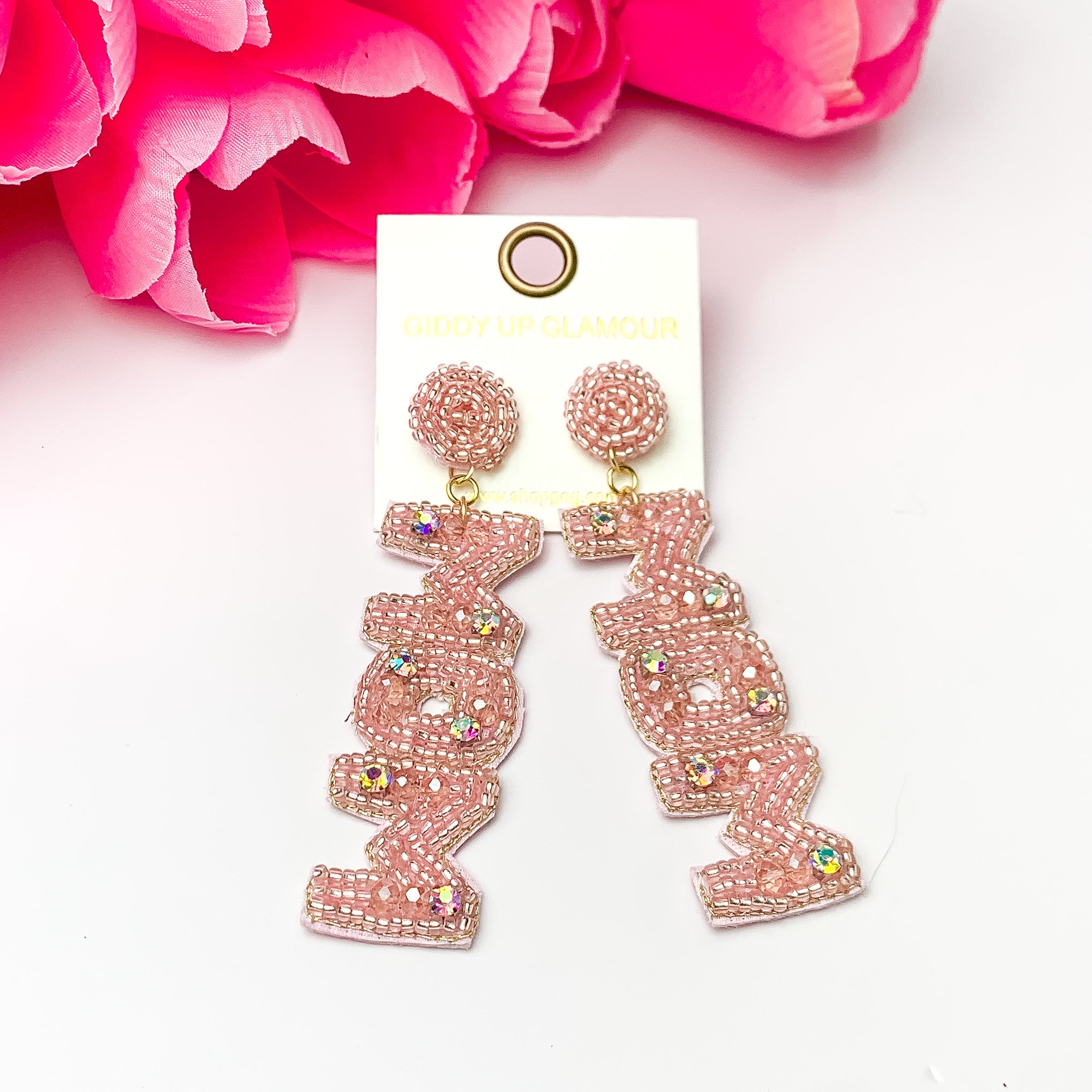 These earrings include rose pink, circle, beaded post back earrings with a long dangle. This dangle spells out "MOM" in rose pink beads with an inlay of ab crystals. These earrings are pictured on a white background with pink flowers.