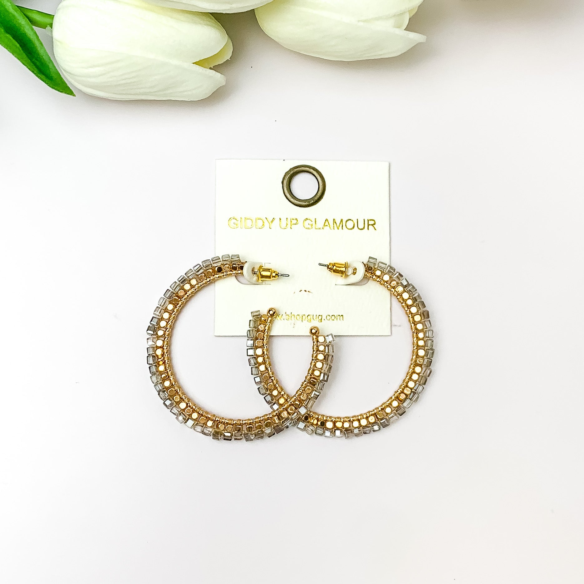  Pictured are circle gold toned hoop earrings with gold beads around it and a grey crystal outline. They are pictured with white flowers on a white background.