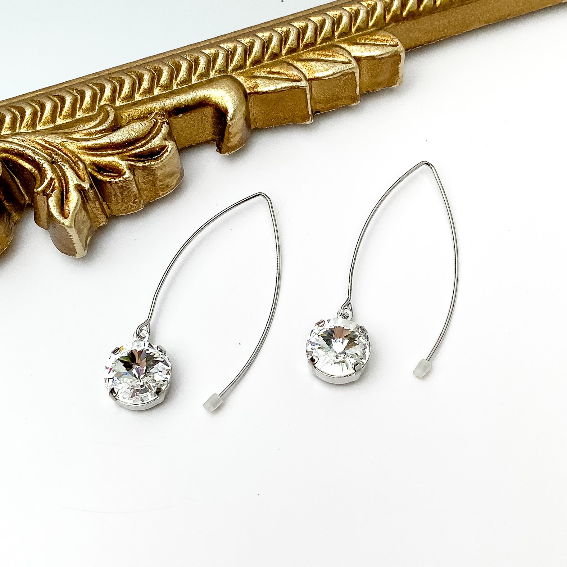 Silver, kidney wire dangle earrings. These earrings include a round clear crystal drop. These earrings are pictured in front of a gold mirror on a white background. 