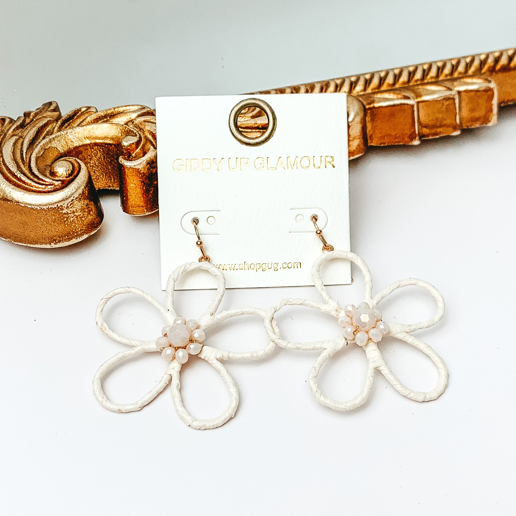 Pictured are gold fish hook earrings with a flower outline pendant. This pendant is ivory and has center ivory crystals. These earrings are pictured in front of a gold mirror on a white background. 