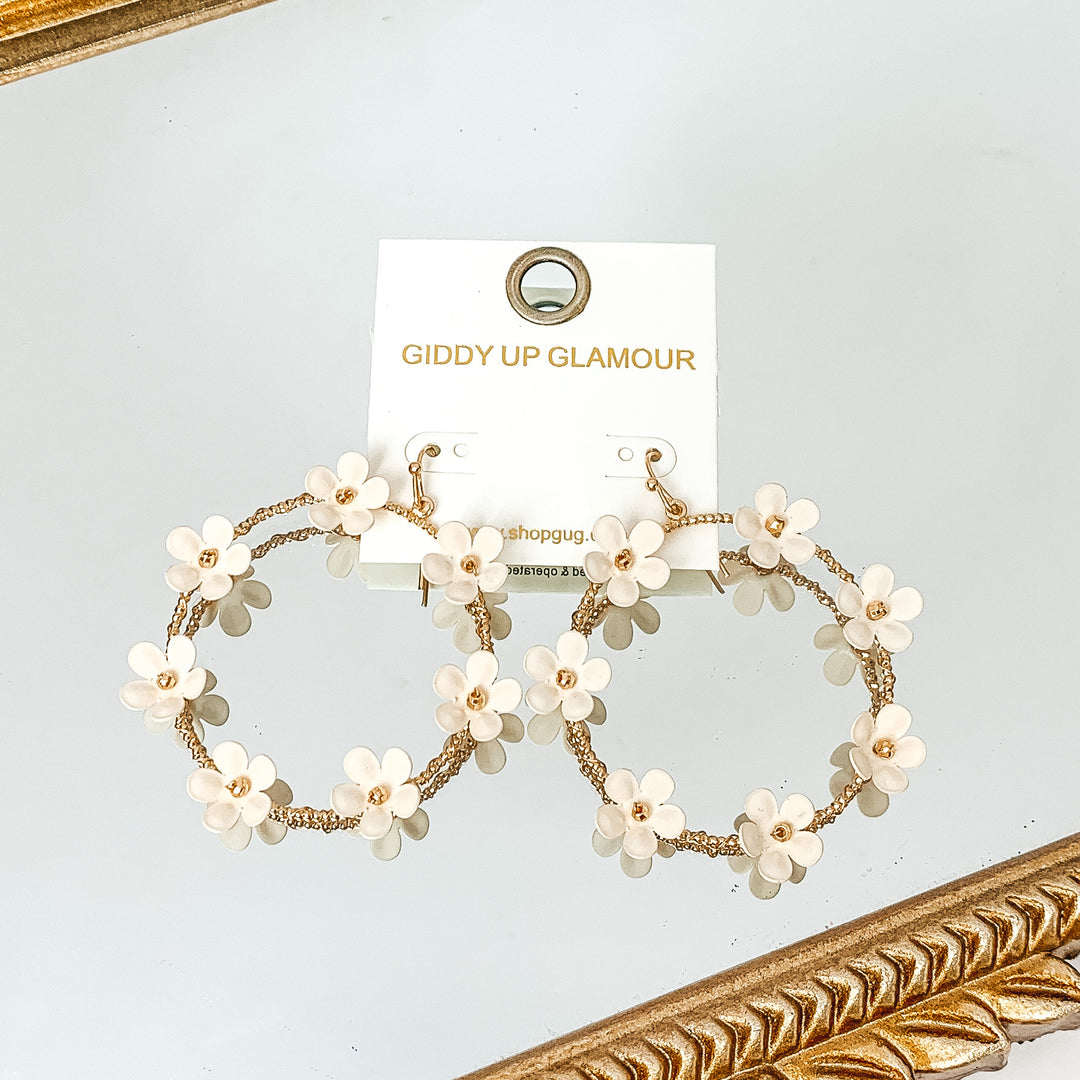 Gold, open circle drop earrings with ivory flower charms outlining the earrings. These flowers have a gold bead center. These earrings are pictured laying on a gold mirror on the white background. 