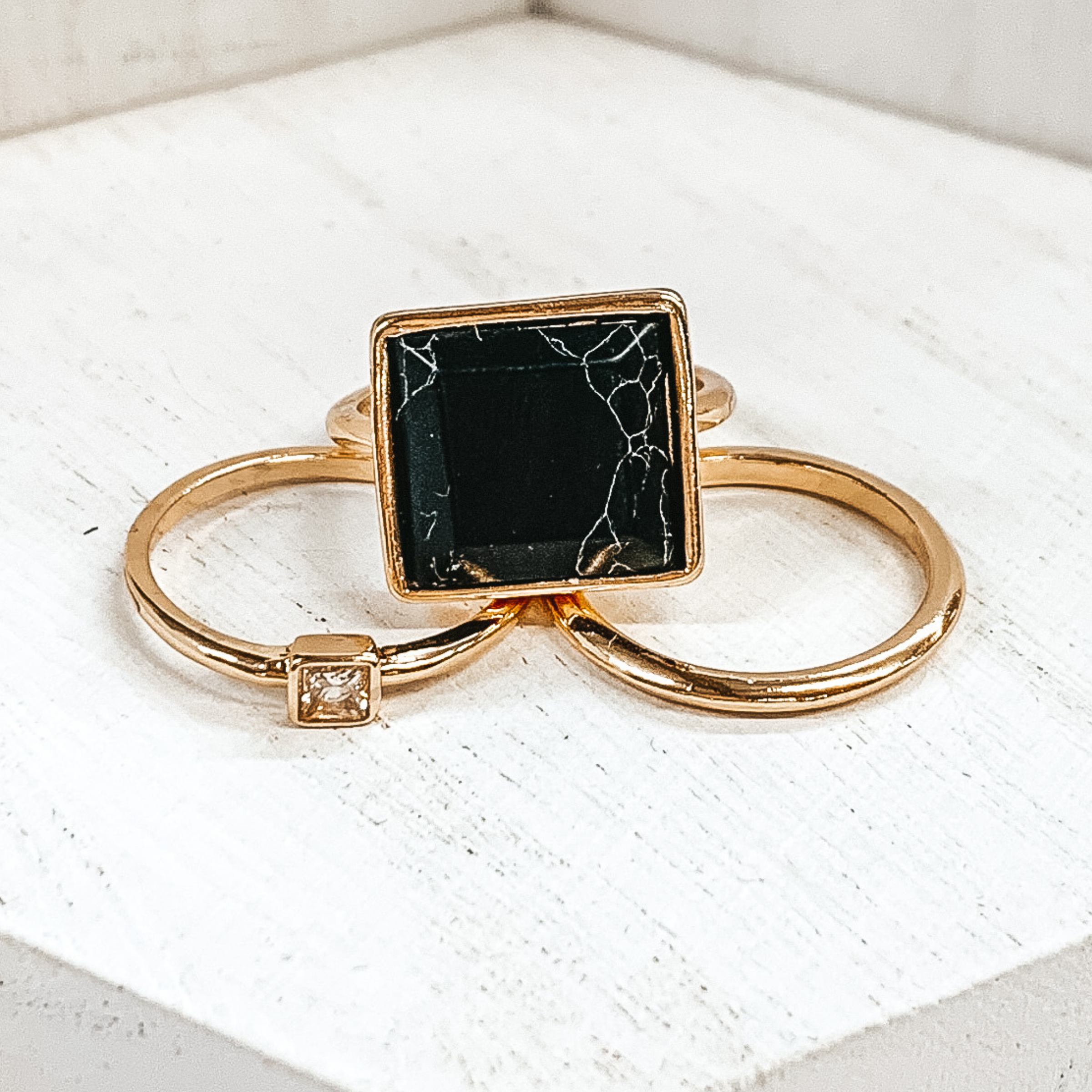 Set of three gold rings. One ring is a plain gold band. Another has a small square pendant with a center crystal. The third ring has a big, black pendant. These rings are pictured on a white background.  