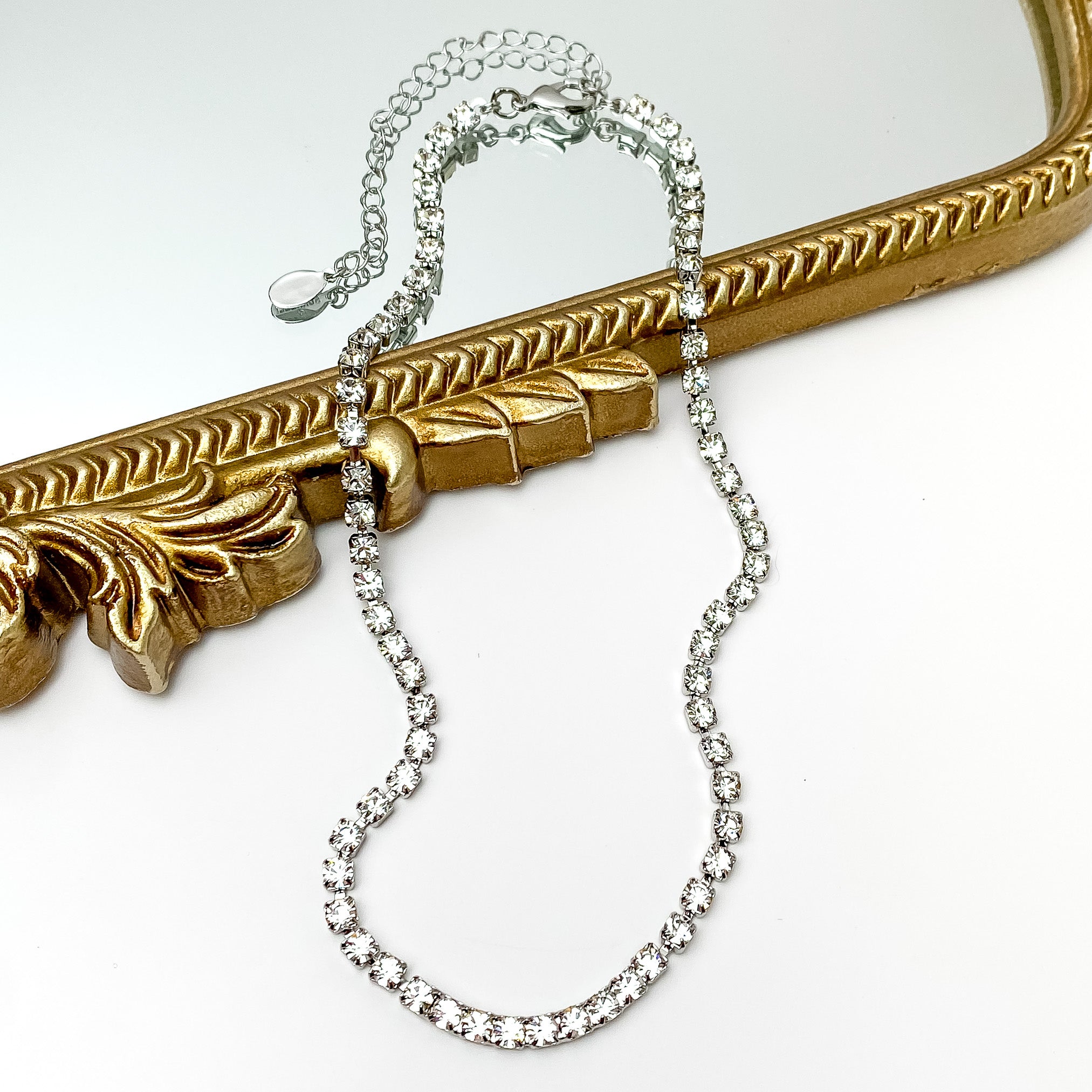 Pictured is a silver necklace with clear crystals. This necklace is pictured partially on a gold mirror on a white background. 