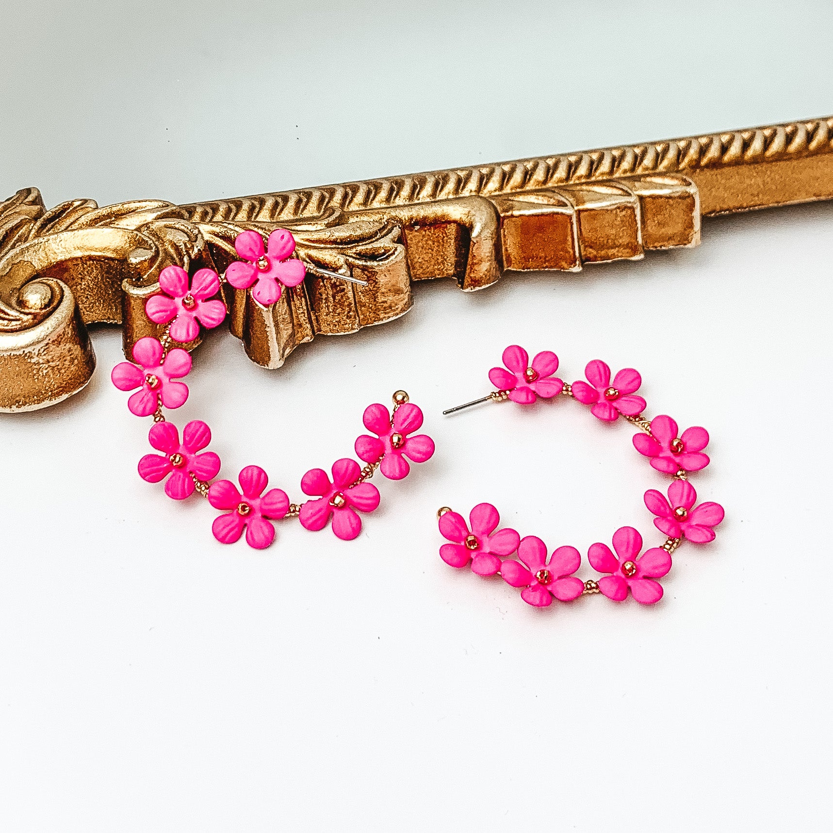 Gold hoop earrings with hot pink flower charms outlining the earrings. These flowers have a gold bead center. One hoop is pictured laying against a gold mirror while the other hoop is laying flat on the white background. 