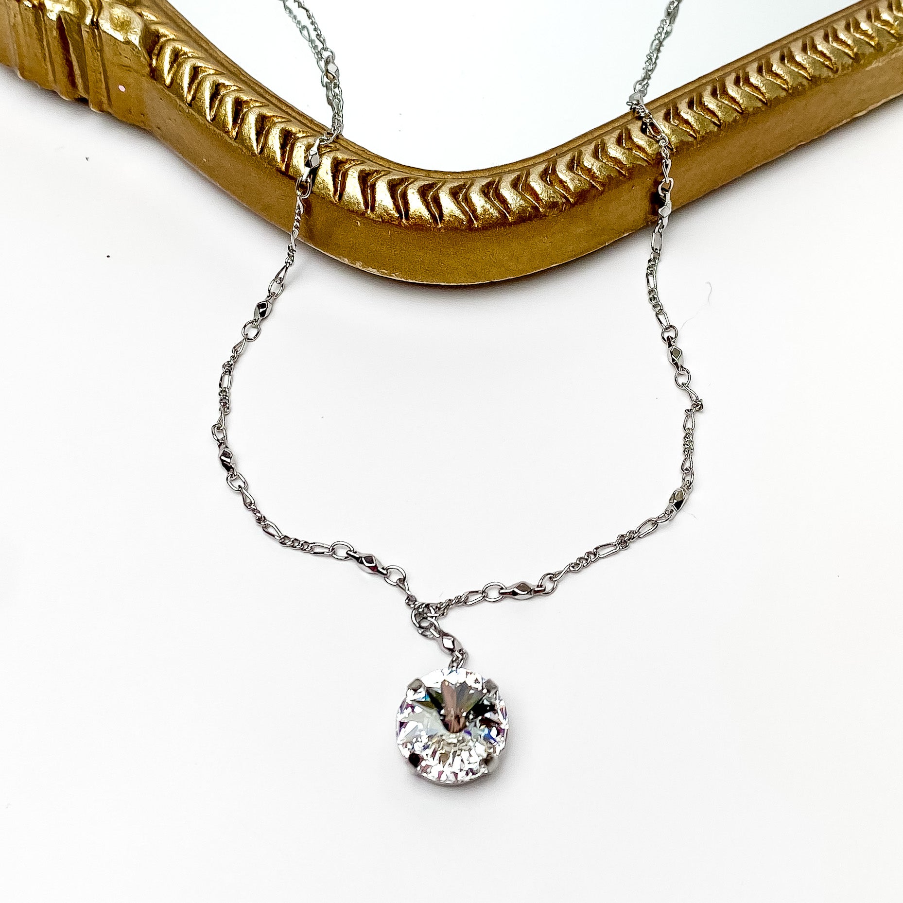 Pictured is a silver necklace with a clear crystal pendant. This necklace is pictured laying partially on a gold mirror on a white background. 