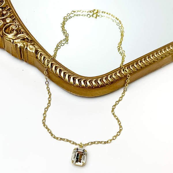 Pictured is a thin, gold chain necklace with a single, emerald cut clear crystal. This necklace is pictured partially laying on a gold mirror on a white background.    