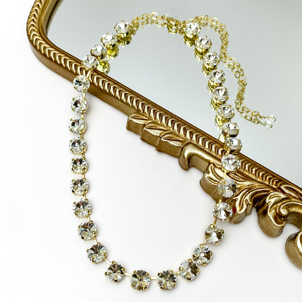 Clear round crystal necklace. This necklace has a gold setting. This necklace is pictured partially laying on a gold mirror on a white background.    