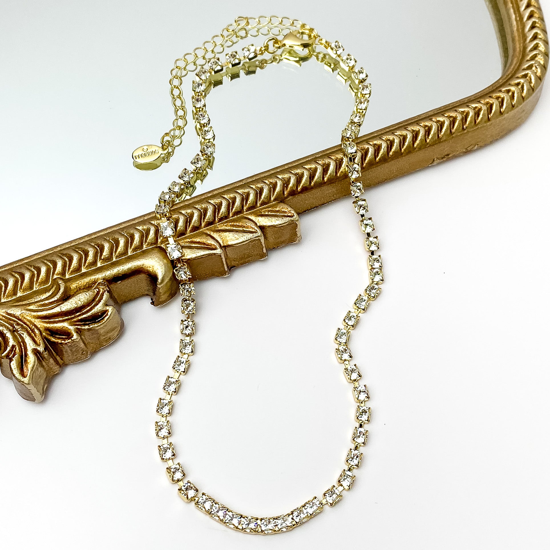 Pictured is a gold necklace with clear crystals. This necklace is pictured partially on a gold mirror on a white background.
