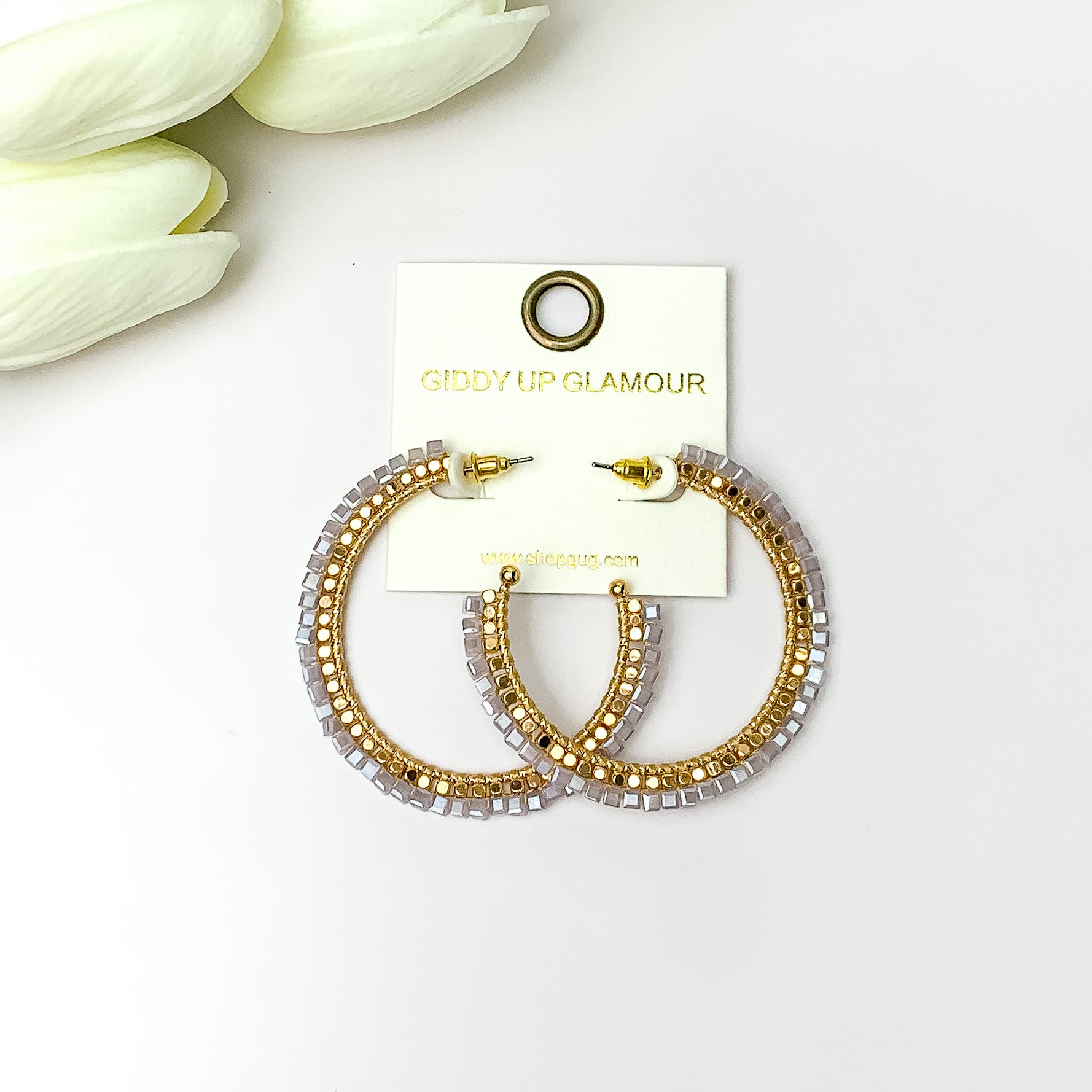 Pictured are circle gold toned hoop earrings with gold beads around it and pale lavender crystal outline. They are pictured with white flowers on a white background.