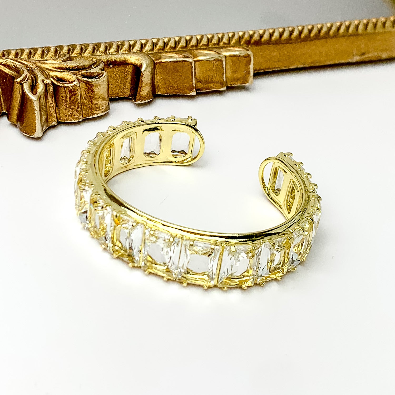 Gold bangle with a clear, rectangle crystal inlay. This bracelet is pictured on a white background with a gold mirror at the top of the picture.  