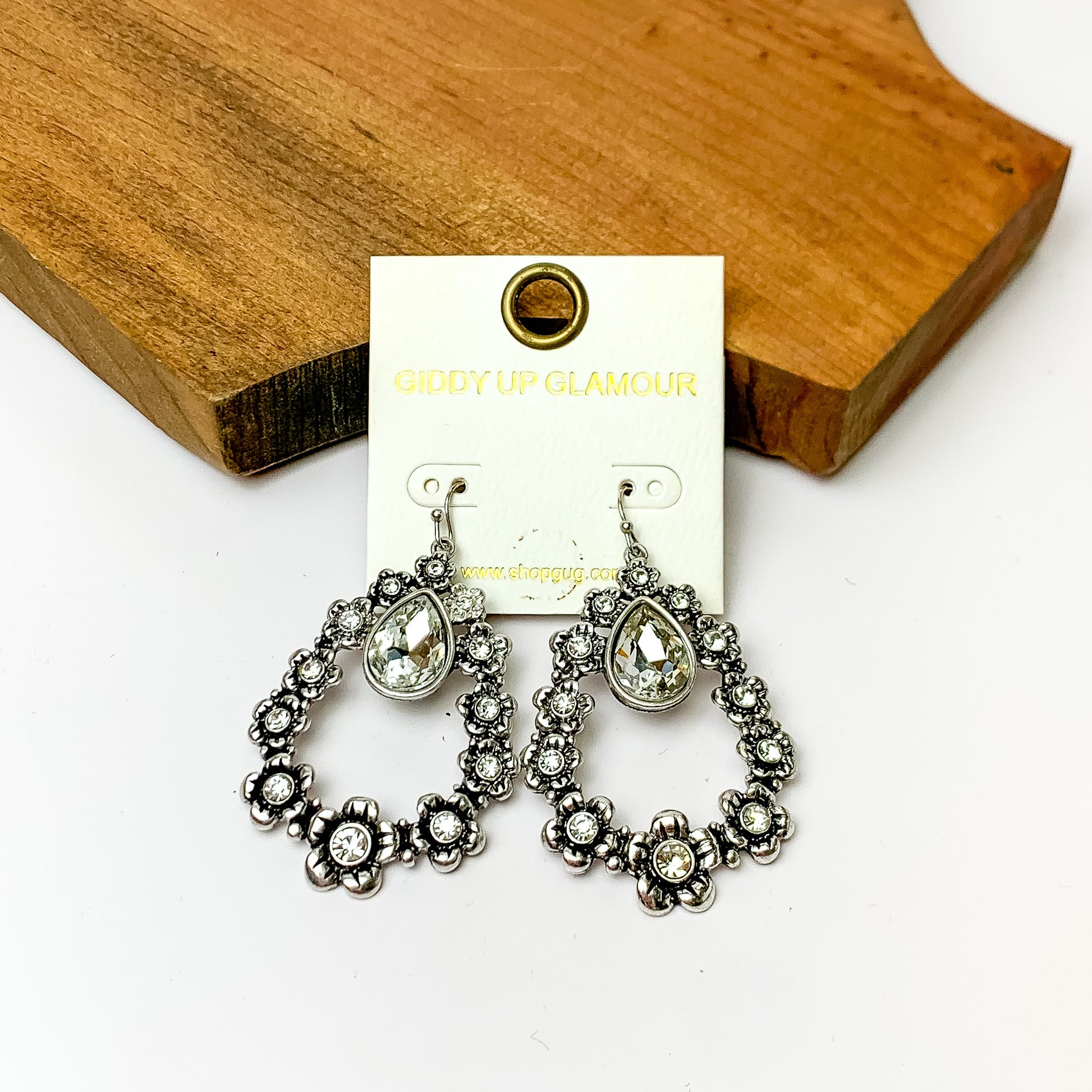 Silver open drop earrings with flowers connecting and clear crystals. Pictured on a white background with a wood piece at the top.
