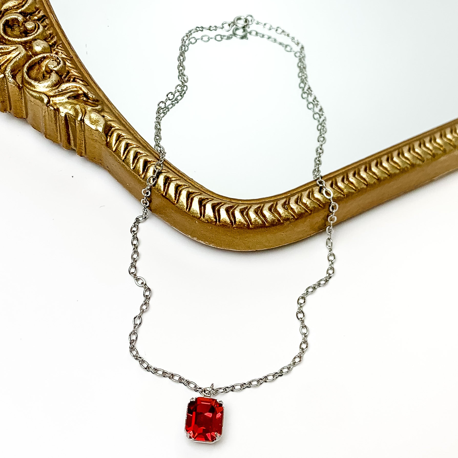 Pictured is a thin, silver chain necklace with a single, emerald cut red crystal. This necklace is pictured partially laying on a gold mirror on a white background.    