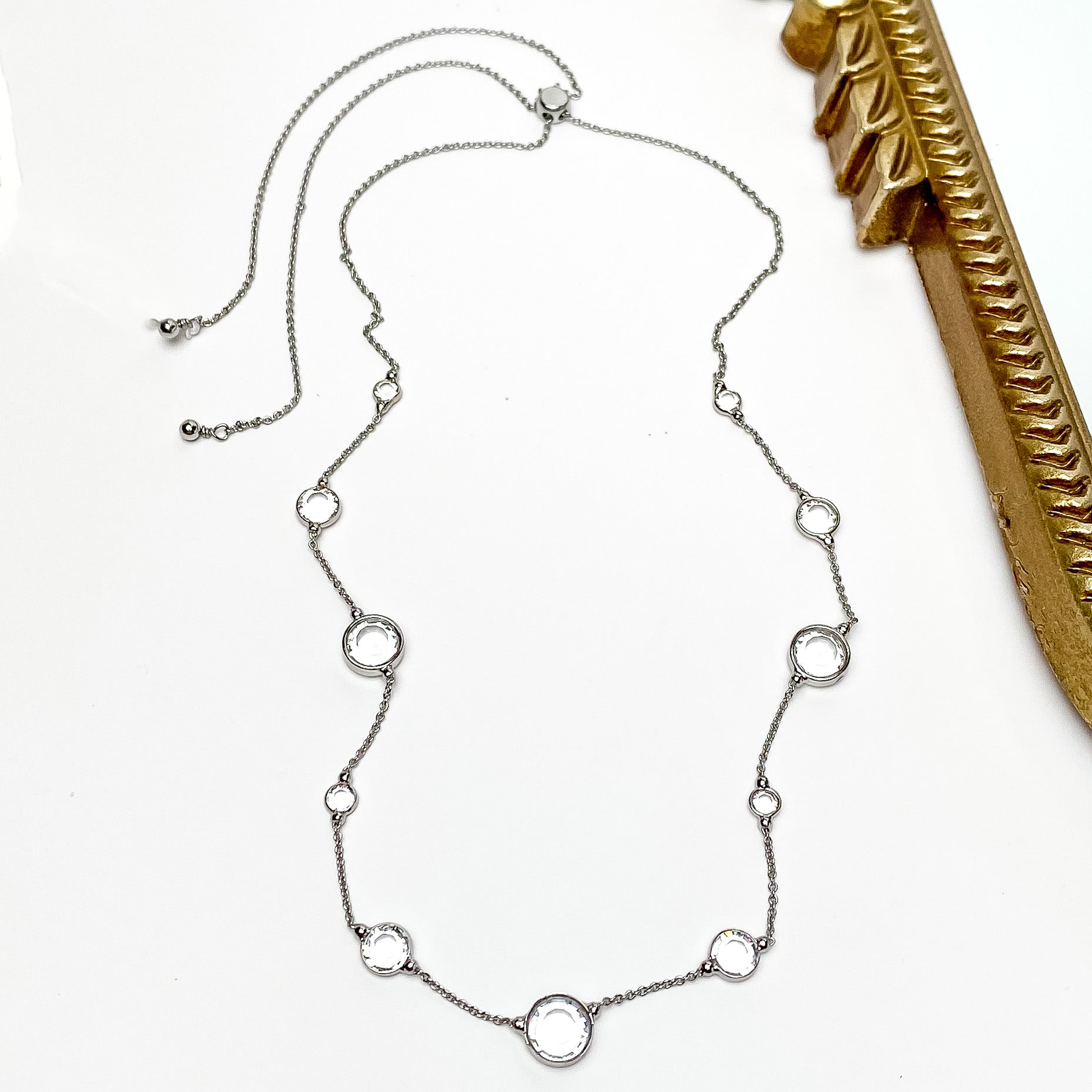 Pictured is a thin, silver chain with spaced out circle clear crystals. These crystals also come in different sizes. This necklace is pictured on a white background with a gold mirror to the right of the necklace.    