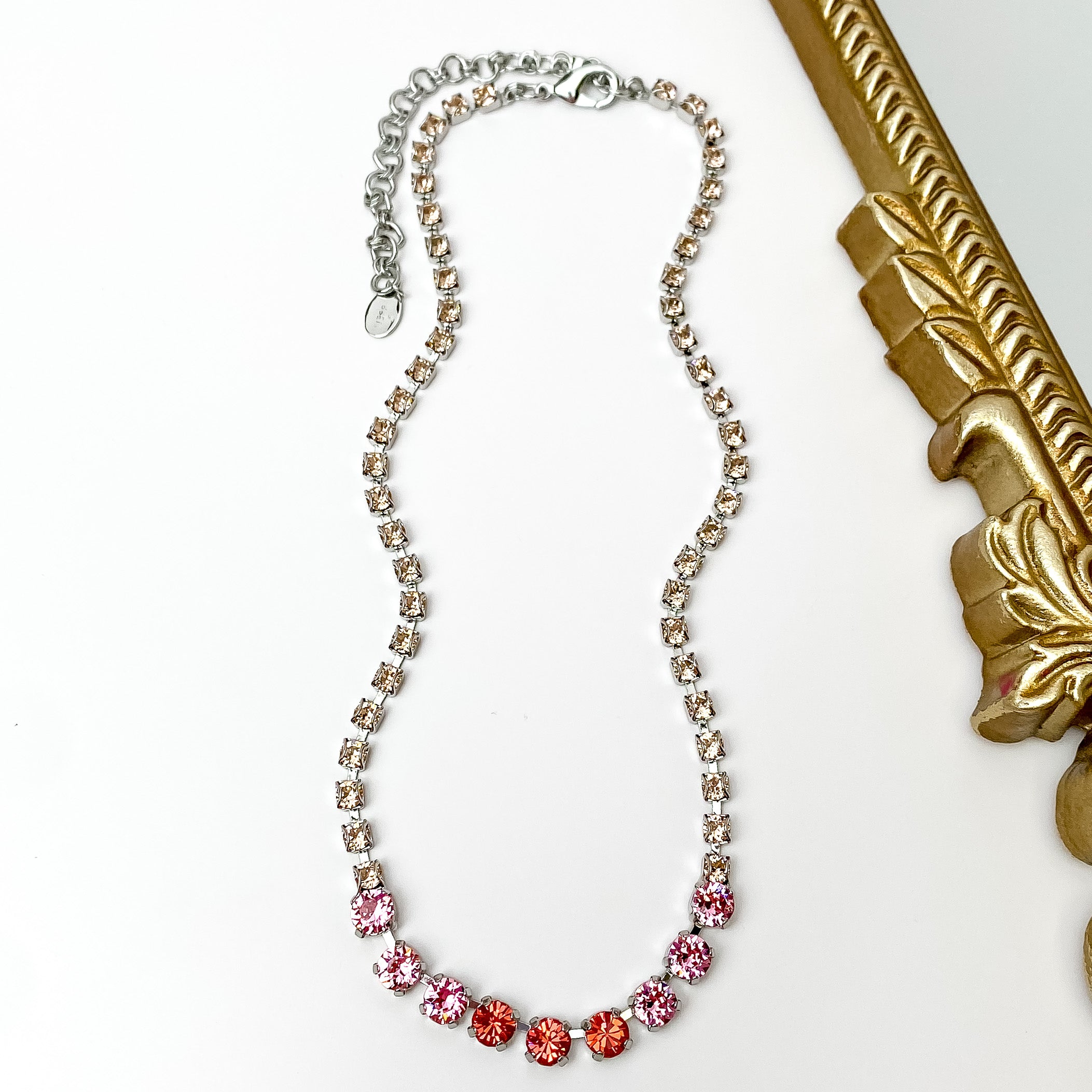 Pictured is a silver necklace with a mix of pink crystals. This necklace is pictured on a white background with a gold mirror on the right side.