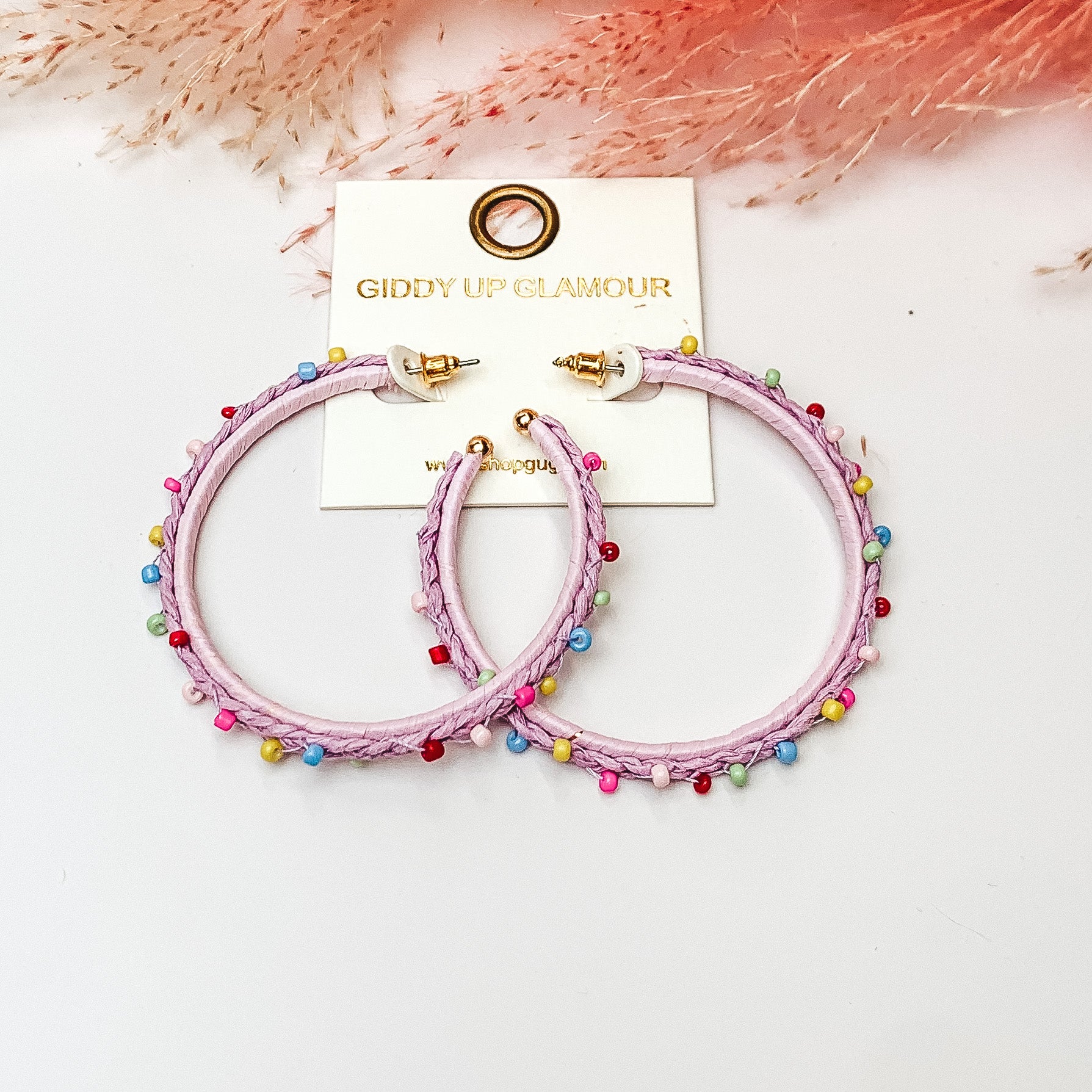 Pictured are raffia braided hoop earrings in lavendar with colorful beads. They are pictured with a pink feather on a white background.