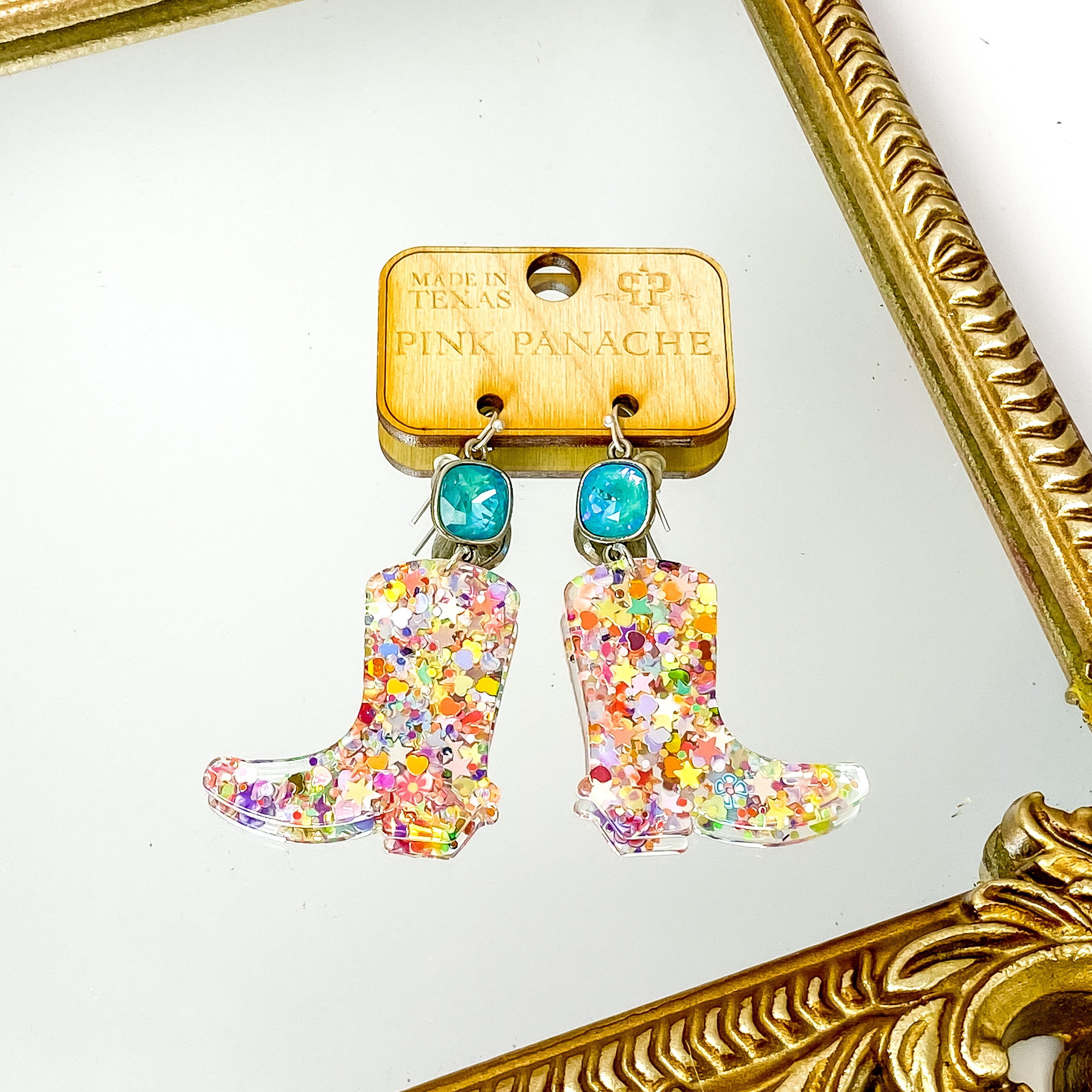 Silver, cushion cut laguna delight crystal drop earrings with a hanging boot pendant. This pendant has multicolor glitter all over. These earrings are pictured on a wood earrings holder on a gold mirror on a white background. 