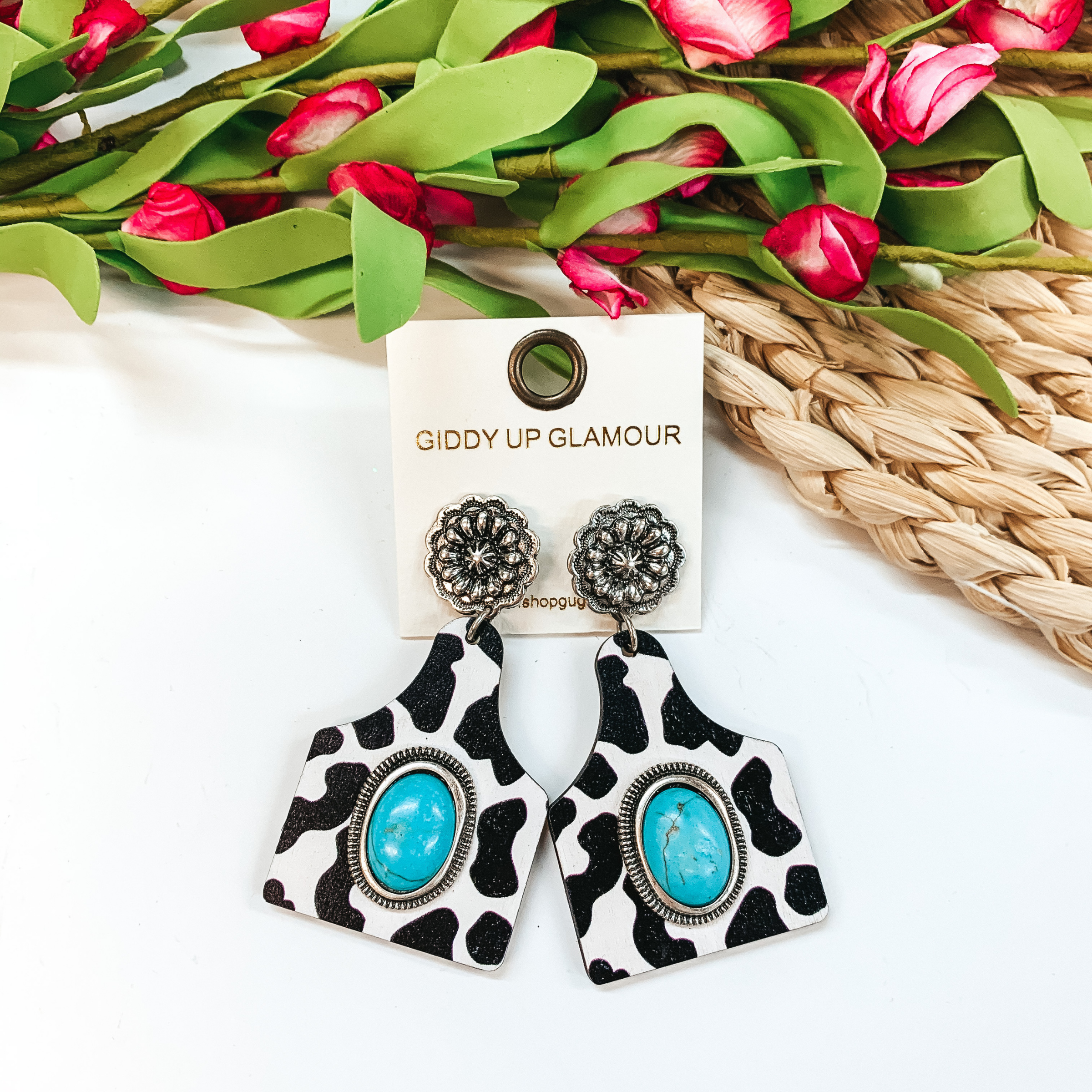 Silver concho flower post back earrings with a hanging cow tag pendant. The pendant has a printed black and white cow print design with a oval turquoise stone. These earrings are pictured on a white background with pink flowers at the top.