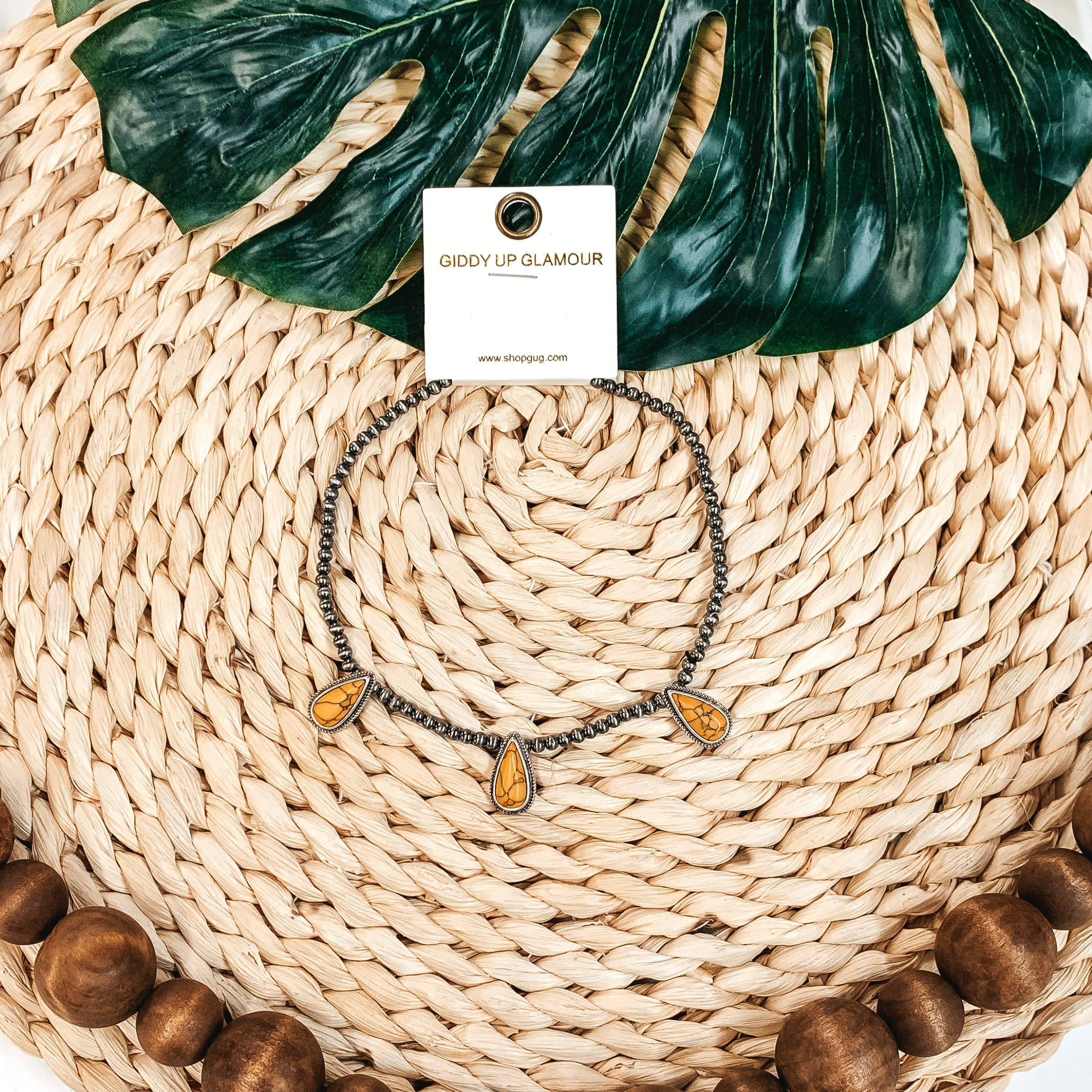 Silver beaded necklace with three yellow, teardrop stone charms. This necklace is pictured on a tan basket weave with a green leaf in the background. 