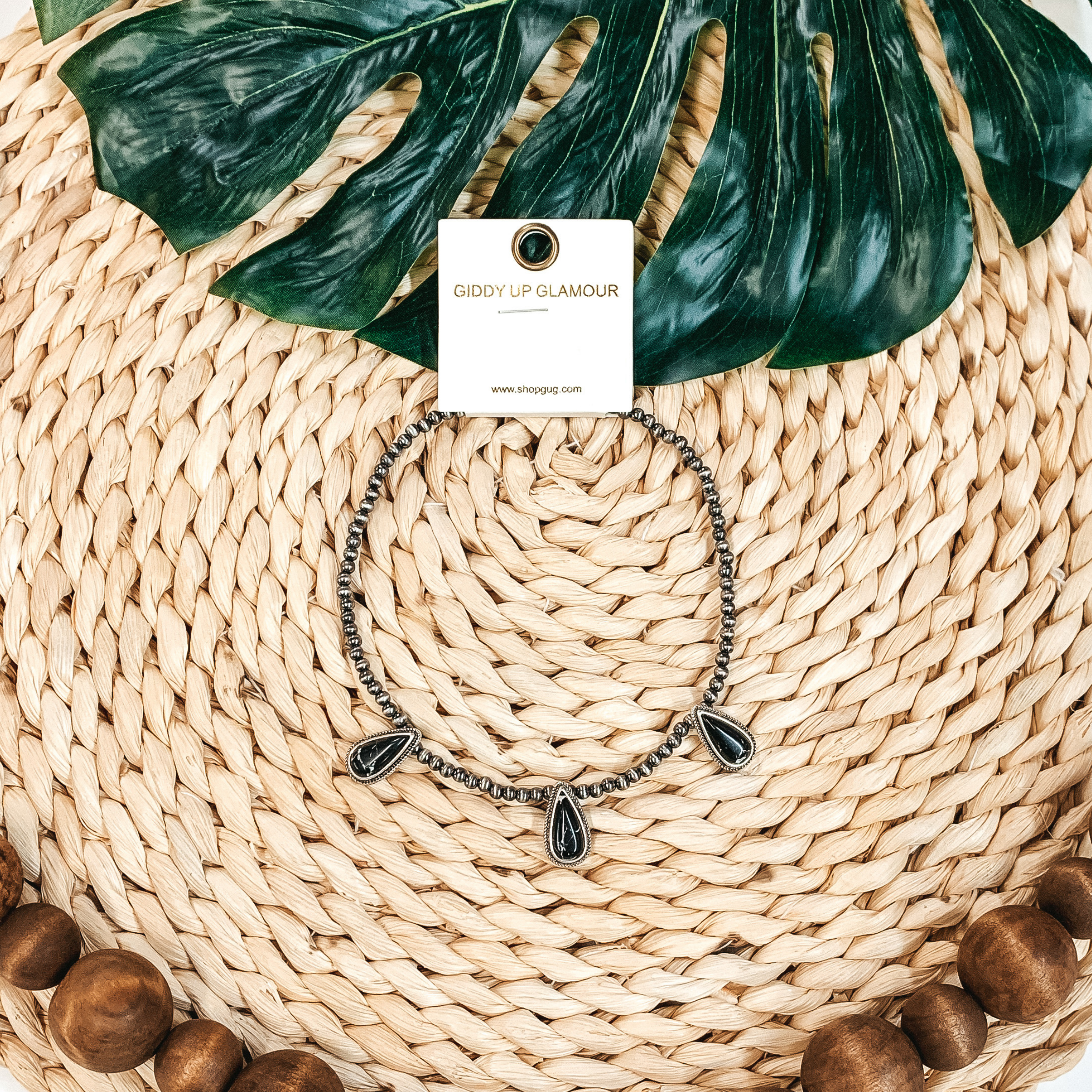 Silver beaded necklace with three black, teardrop stone charms. This necklace is pictured on a tan basket weave with a green leaf in the background.