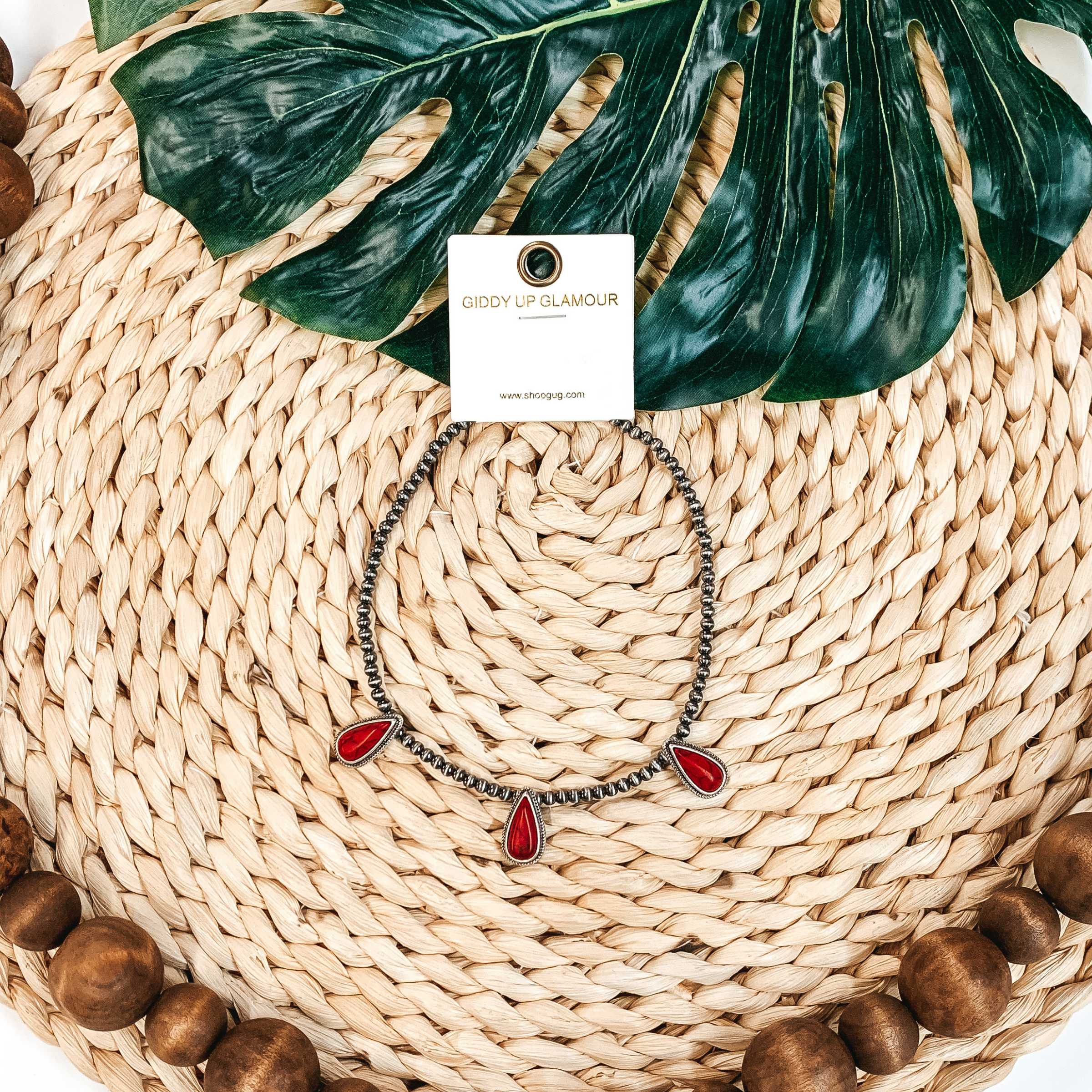 Silver beaded necklace with three red, teardrop stone charms. This necklace is pictured on a tan basket weave with a green leaf in the background.
