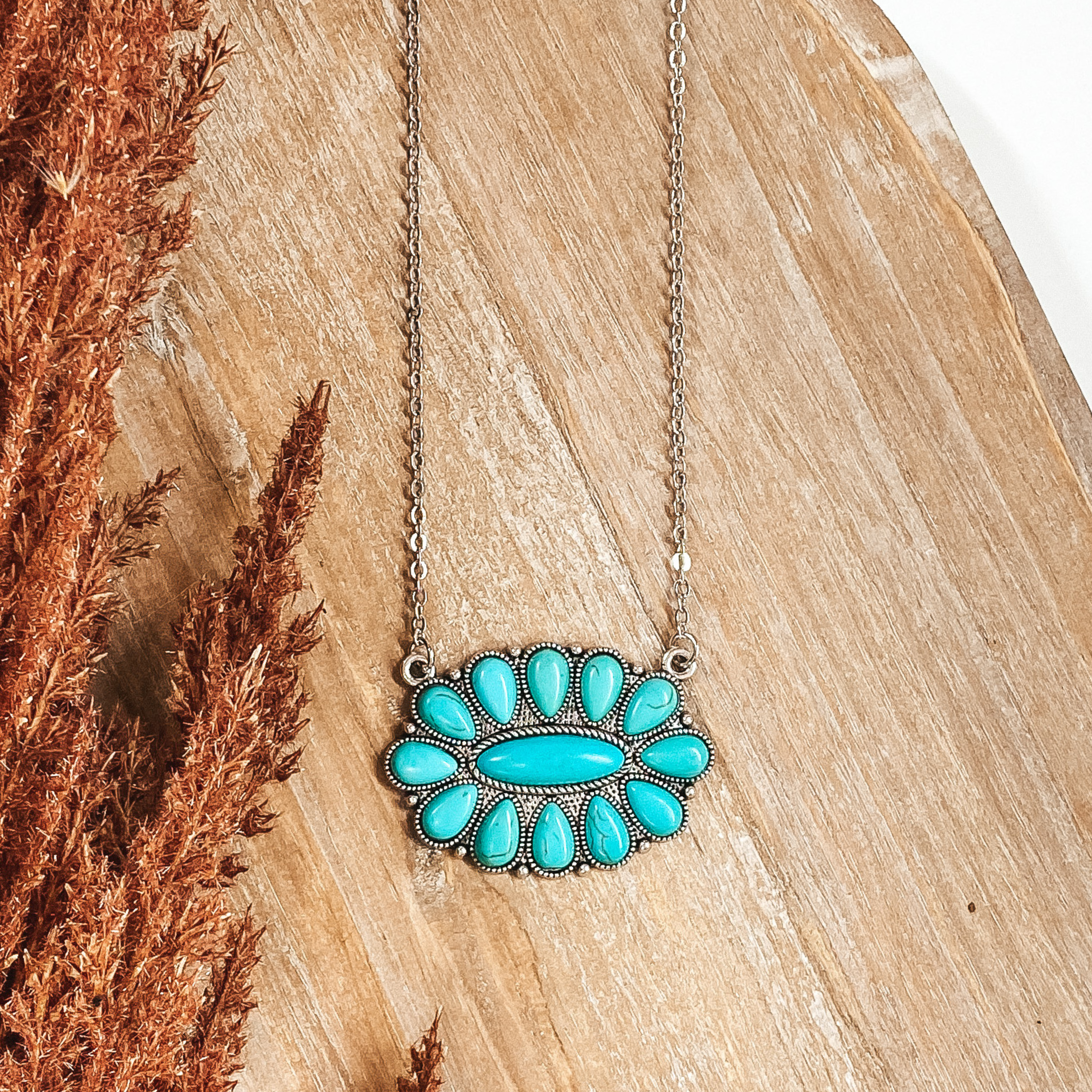 Silver chain necklace with a oval pendant with turquoise stones. This necklace is pictured on a tan background with brown pompous grass lining the left side on a white background.