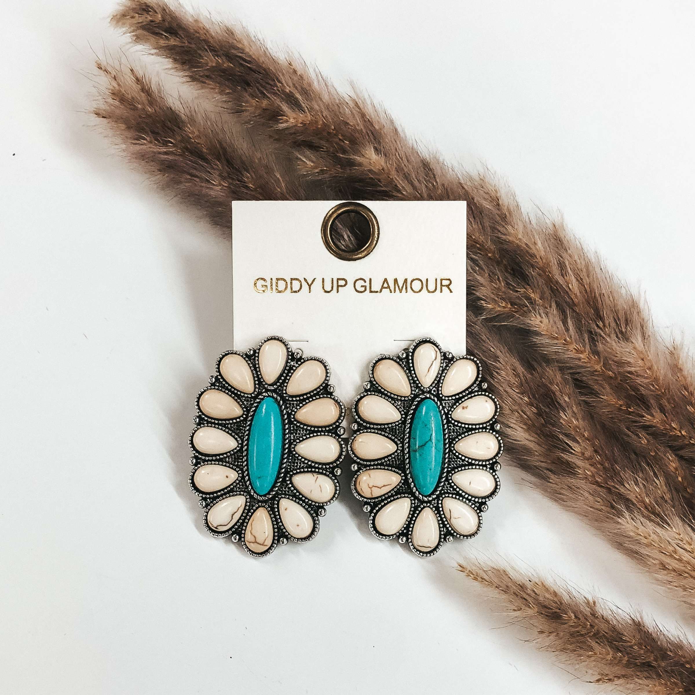 Oval cluster earrings with white stones. These earrings also include a center, oval turquoise stone. These earrings are pictured on a white background in front of brown pompous grass. 