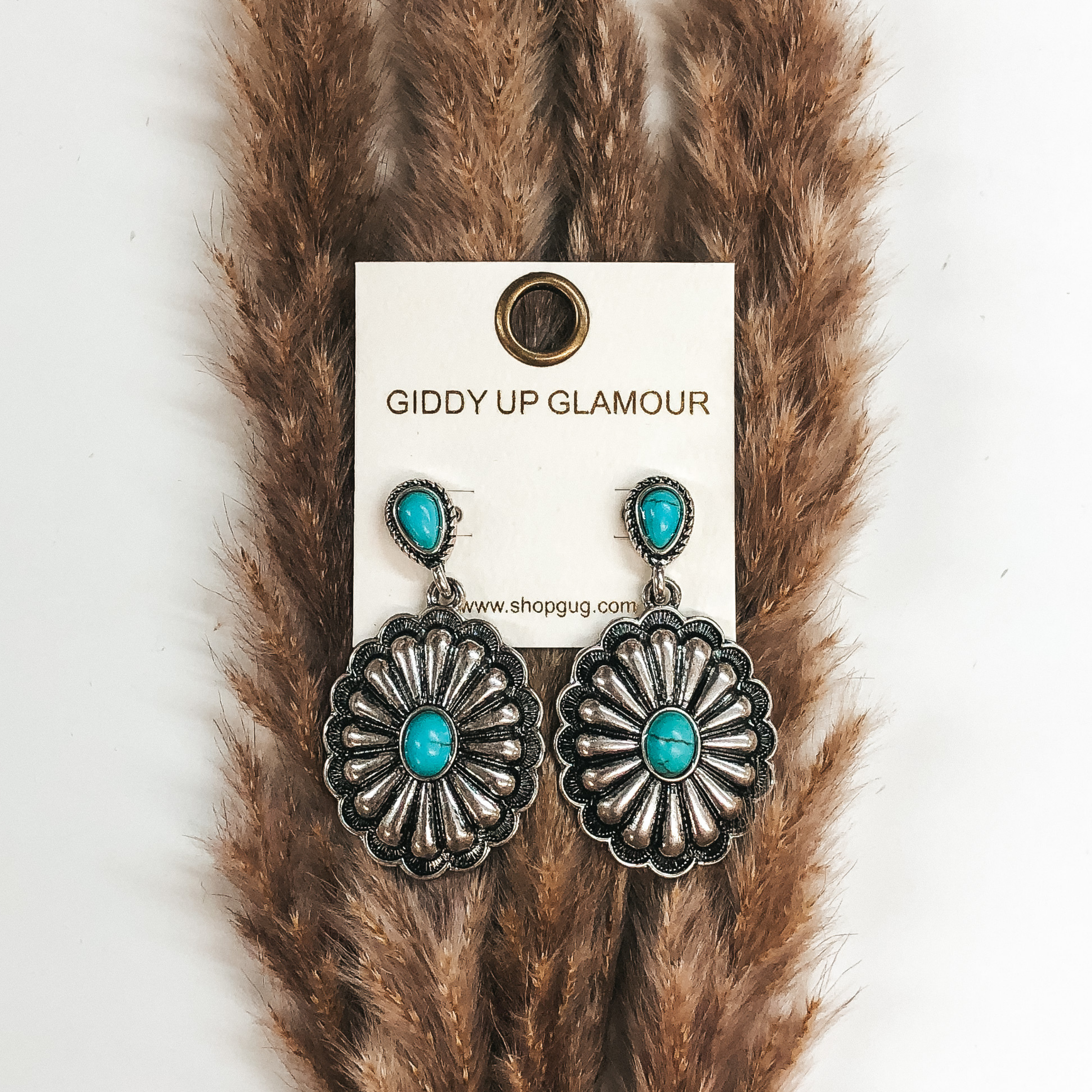 A pair of silver concho earrings with turquoise stones. These earrings are pictured on a white background with a basket and palm leaf.