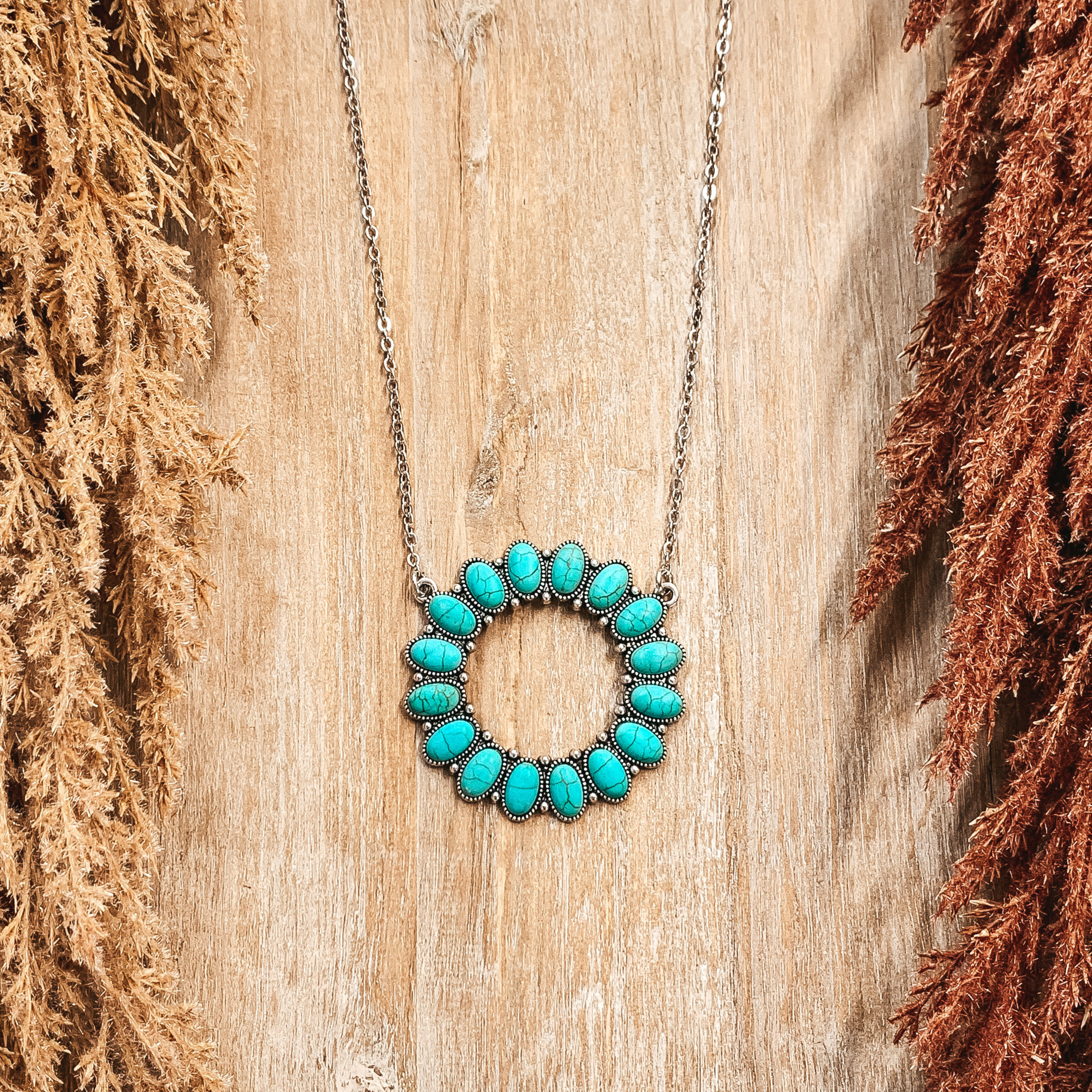 Silver chain necklace with a open circle pendant with turquoise stones. This necklace is pictured on a tan background with brown pompous grass lining the sides. 